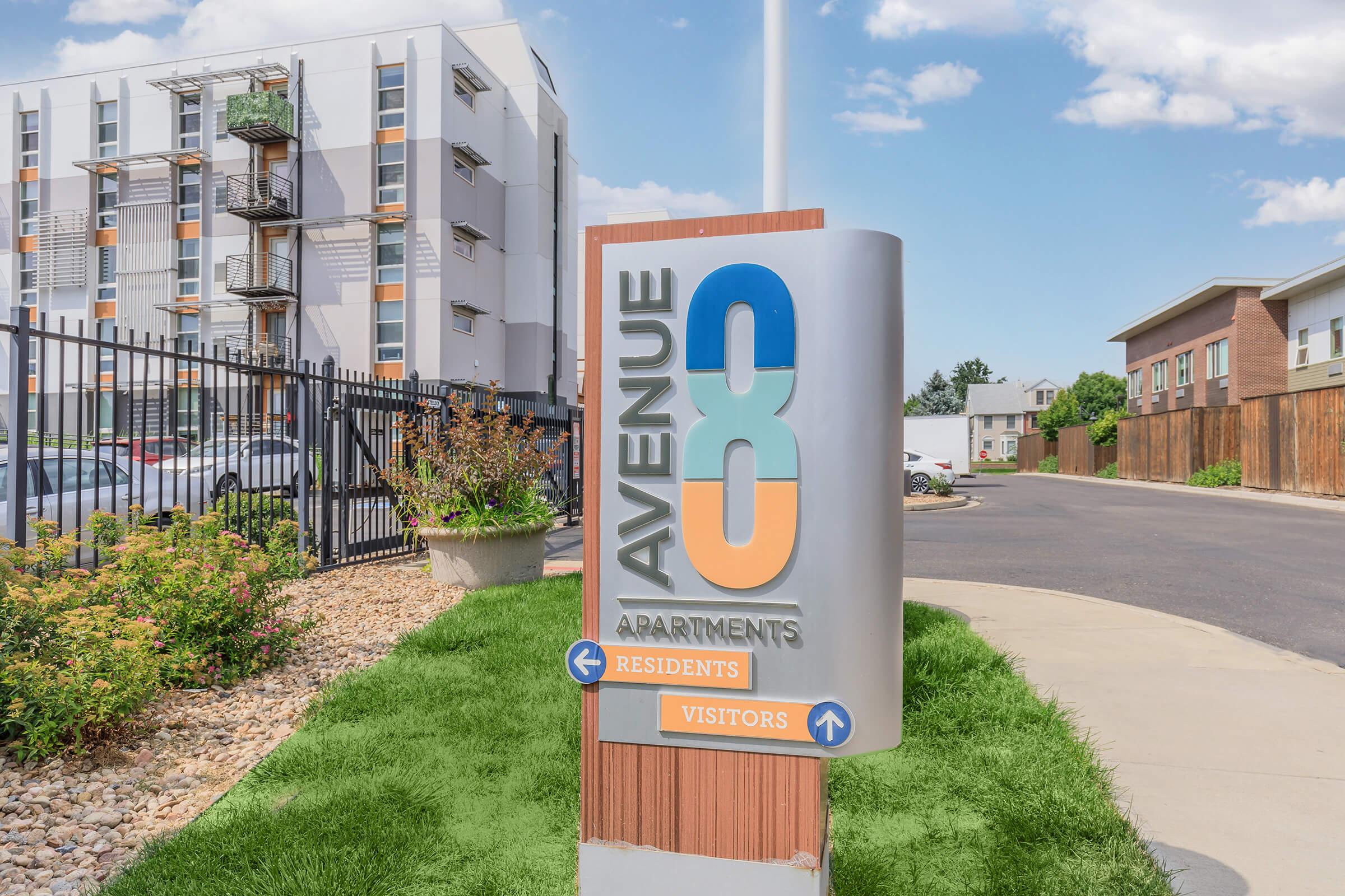 Sign for Avenue 88 Apartments, displaying the words "Residents" and "Visitors" with directional arrows. In the background, modern multi-story apartment buildings and a neatly landscaped area can be seen. The setting is bright and sunny, indicating a pleasant atmosphere.