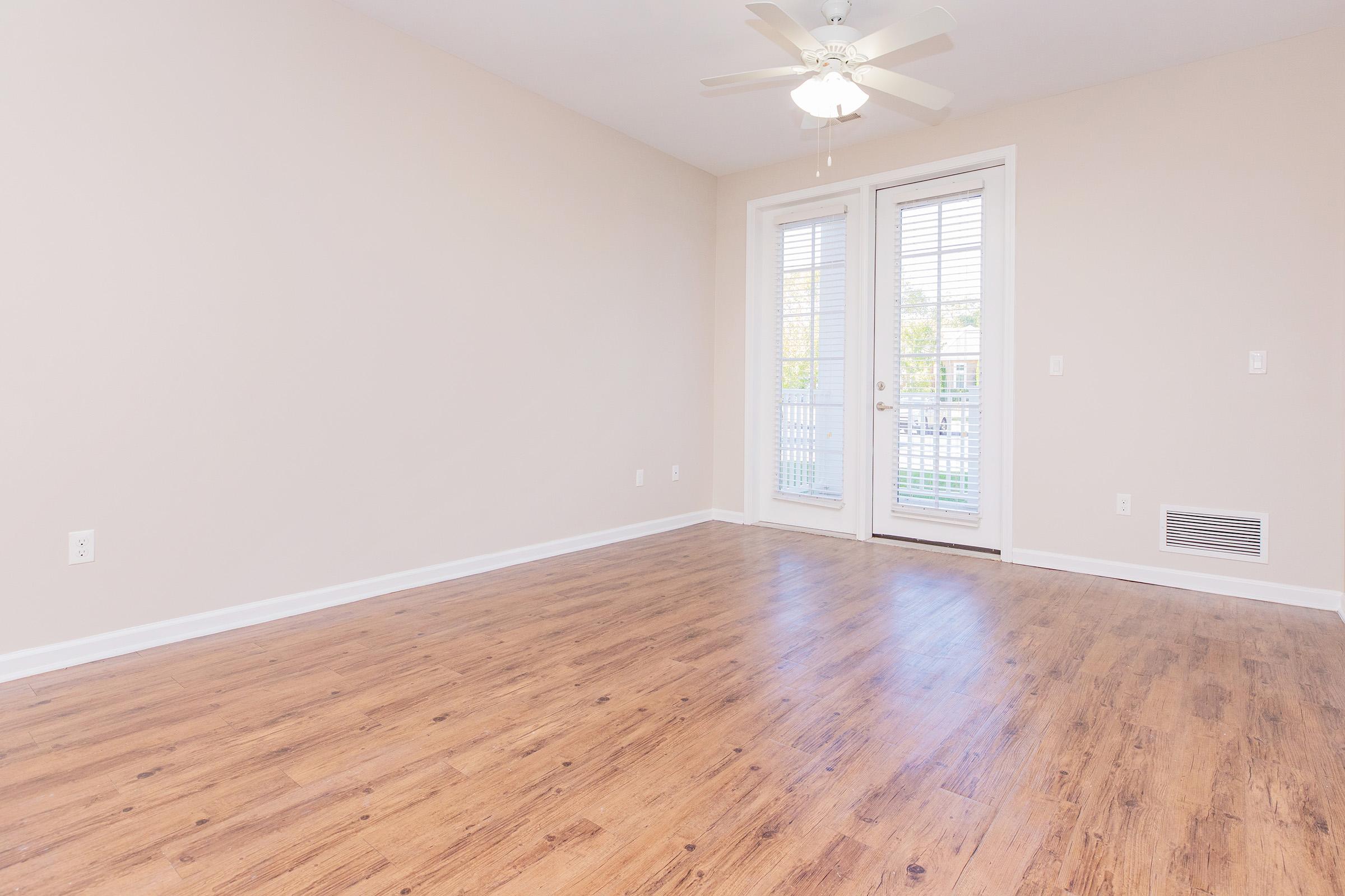 A spacious, empty room with light-colored walls and wooden flooring. It features a ceiling fan and large double doors leading to a balcony with blinds. Natural light floods the space, creating a bright and airy atmosphere.