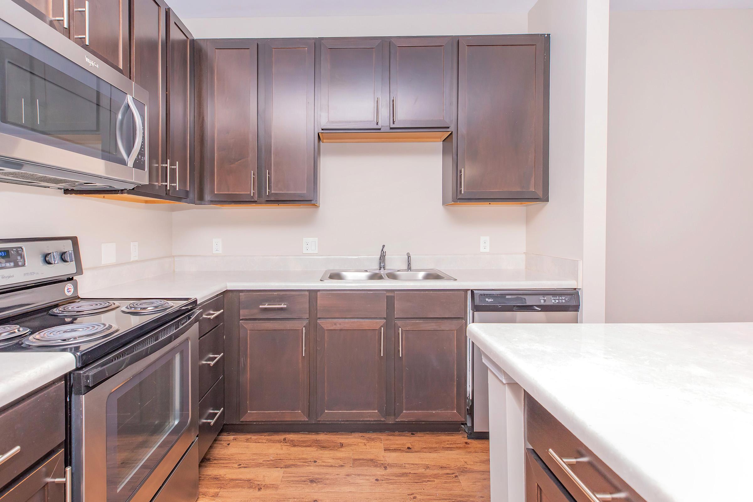 A modern kitchen featuring dark wooden cabinetry, stainless steel appliances including an oven and microwave, a double sink, and a dishwasher. The countertop is light-colored with a smooth finish, and the flooring is a warm-toned wood. The overall design is sleek and contemporary.