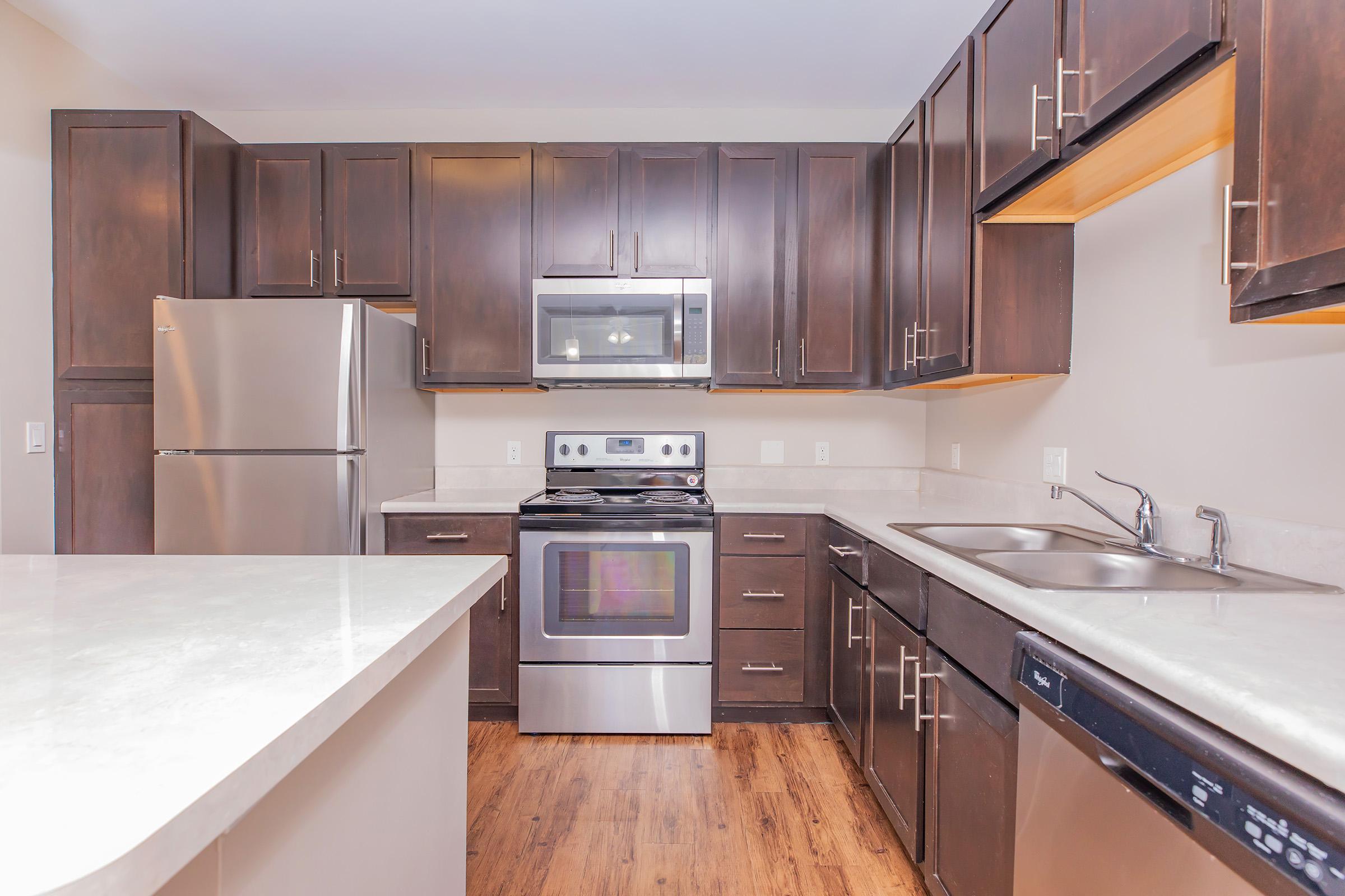 Modern kitchen featuring dark wood cabinetry, stainless steel appliances including a refrigerator and oven, and a light-colored countertop. The kitchen has an open layout with dual sinks and a dishwasher, complemented by wooden flooring and neutral wall colors.