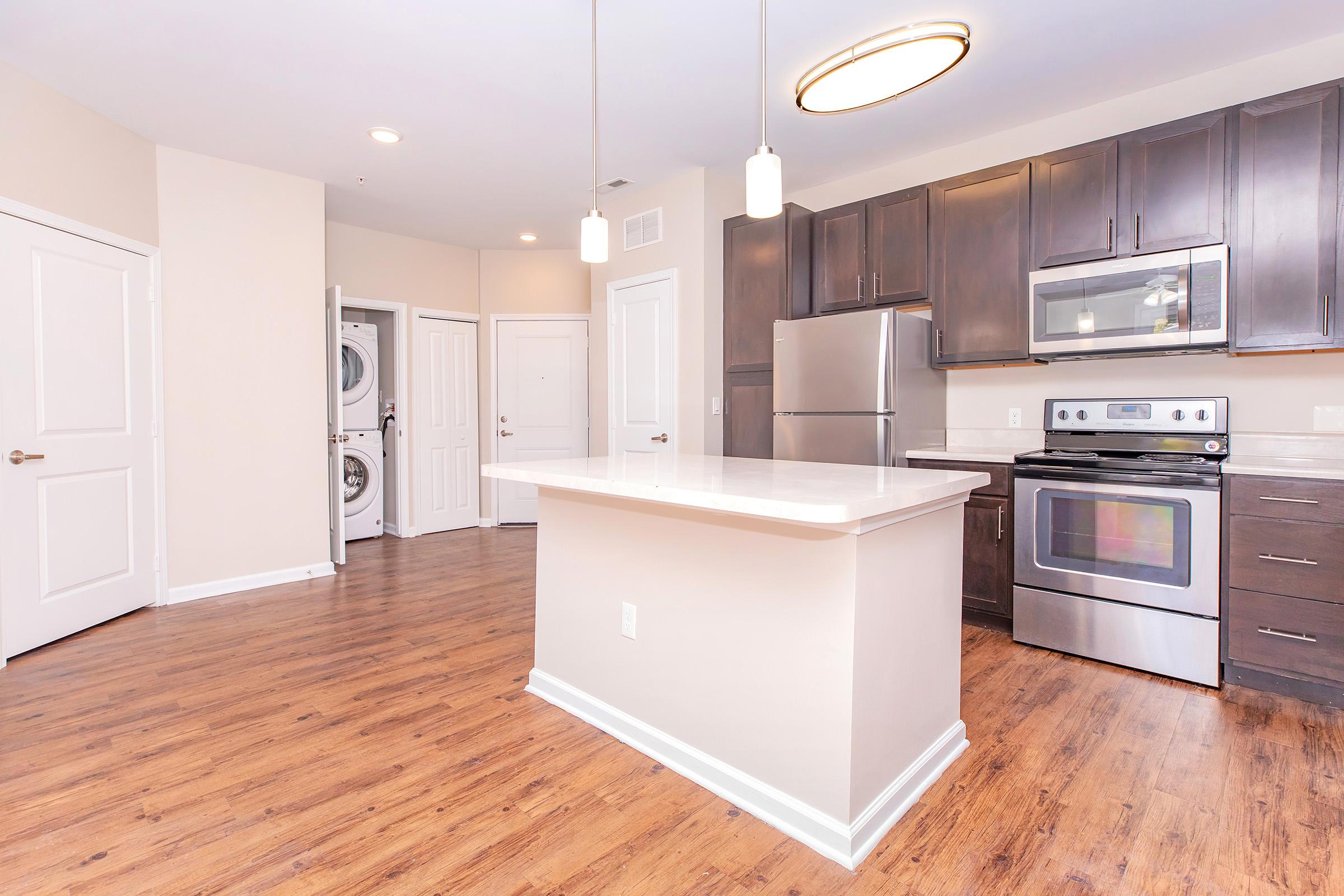 Modern kitchen featuring dark wood cabinetry, stainless steel appliances, and a large island with a white countertop. The open layout includes a nearby laundry area and a door leading to another room. Bright lighting from pendant fixtures enhances the spacious feel with warm wood flooring.