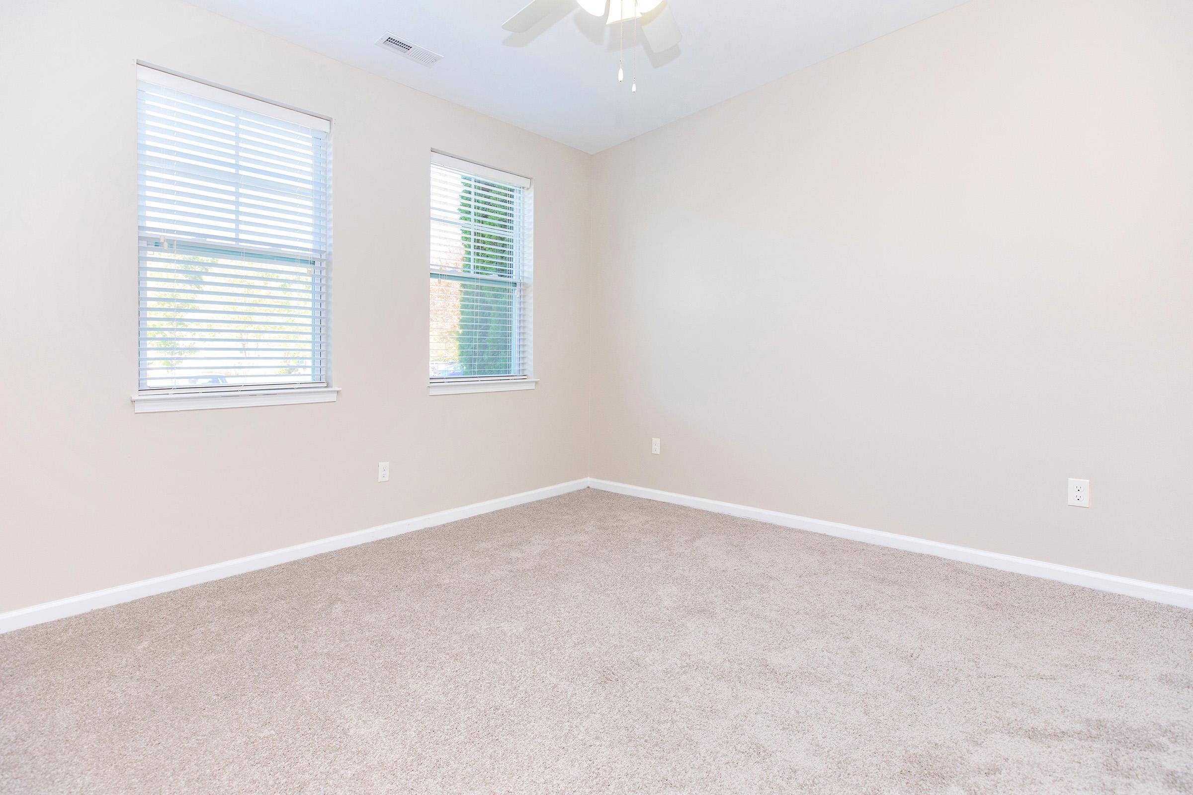 Empty room with beige walls, light-colored carpet, and two windows that let in natural light. A ceiling fan is visible, and there are no furnishings or decorations in the space, creating a clean and minimalist atmosphere.