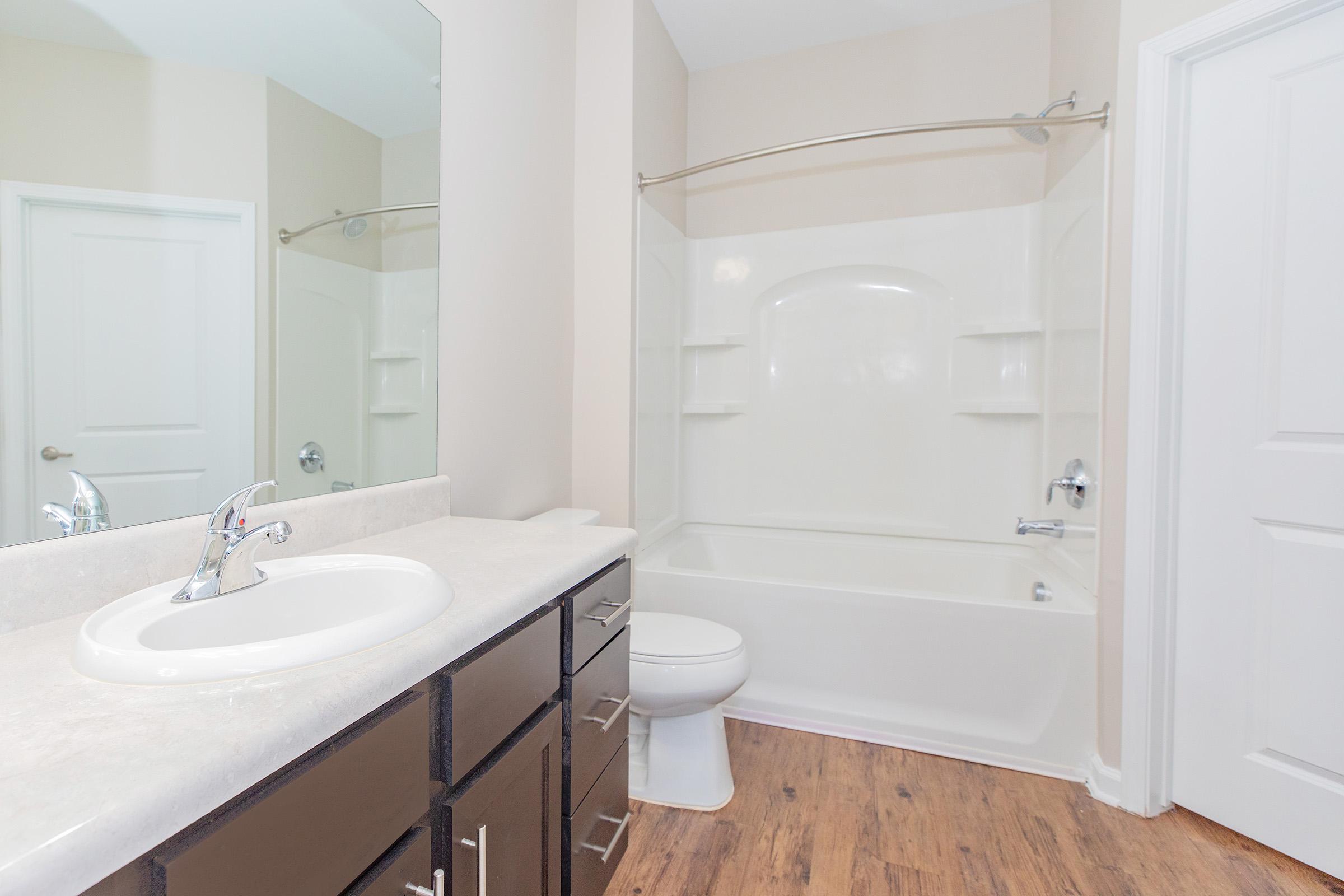 A clean and modern bathroom featuring a white bathtub with built-in shelves, a single sink vanity with a chrome faucet, a large mirror, and a white toilet. The walls are a light beige color, and the flooring is a warm wood finish.