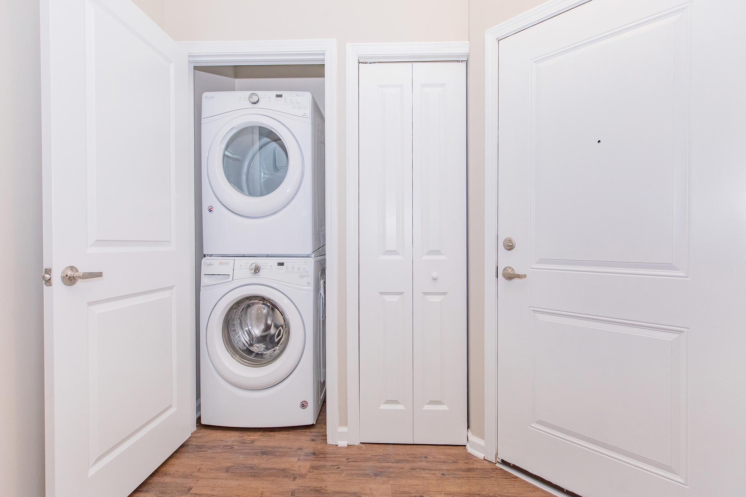 A laundry area featuring a stacked washer and dryer in a narrow space between two white doors. The walls are painted a light color, and the floor is made of wood. One door leads to a closet, while the other appears to be the main entrance.