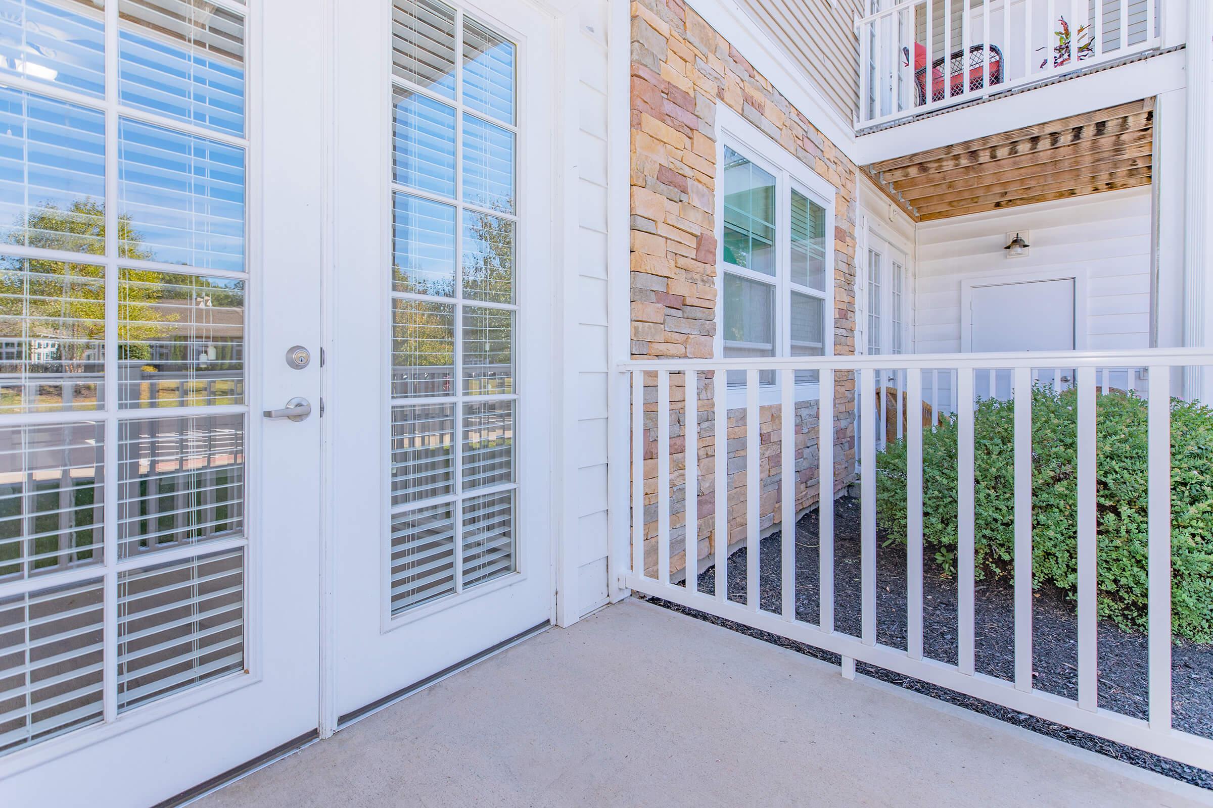 A well-lit entrance featuring double glass doors with white blinds, flanked by stone and wooden textures. A railing surrounds the small porch area, leading to a neatly maintained bush and a glimpse of an adjacent building with outdoor space above.