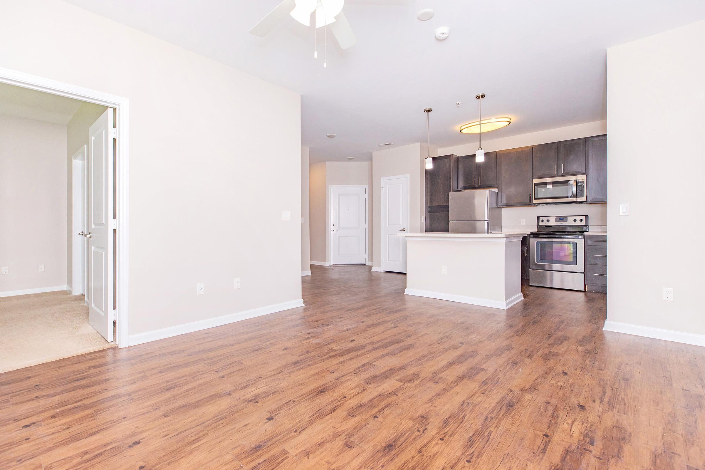 Interior view of a modern apartment featuring an open floor plan. The room has laminate flooring, a ceiling fan, and a kitchen with stainless steel appliances and dark cabinetry. There are two doorways leading to other rooms, and ample natural light fills the space through the large windows.