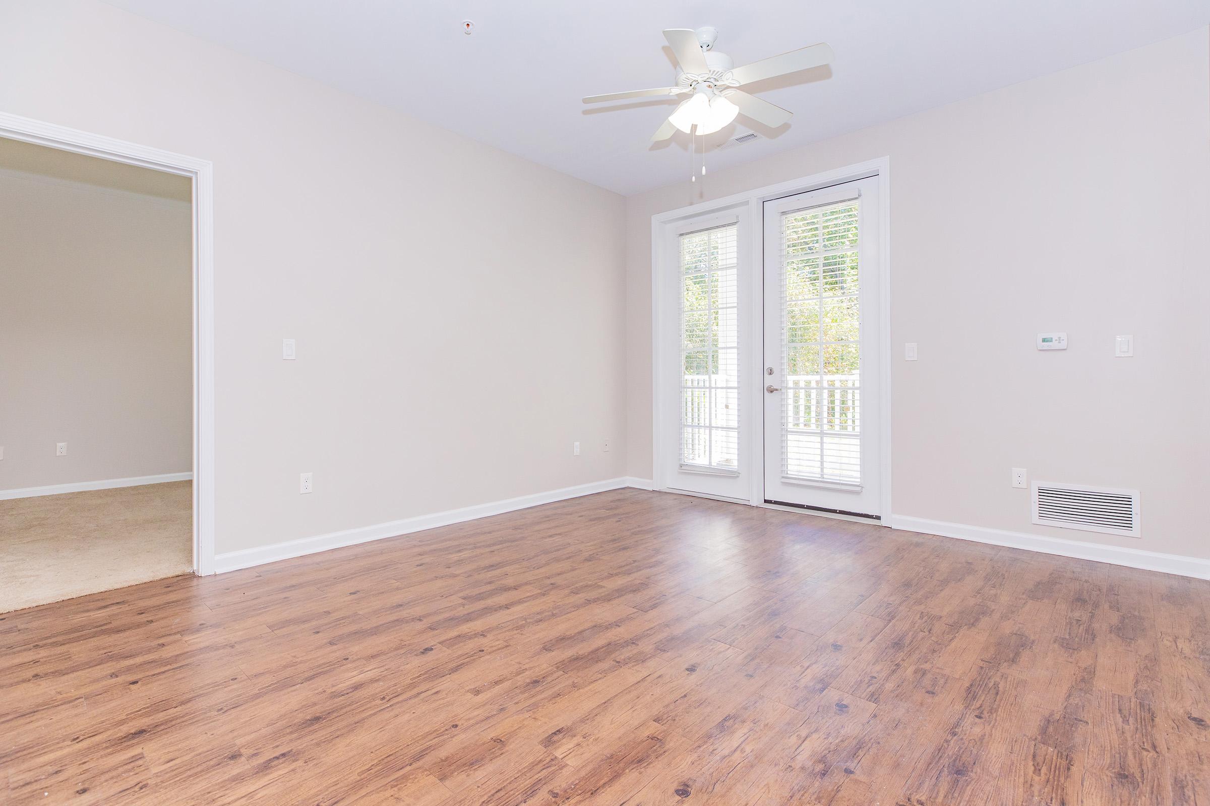 Empty room with light-colored walls and a wooden floor. A ceiling fan is visible, and there are large sliding glass doors leading to a balcony. The opposite wall has an opening to another room. The space is well-lit and inviting, with a neutral color palette.