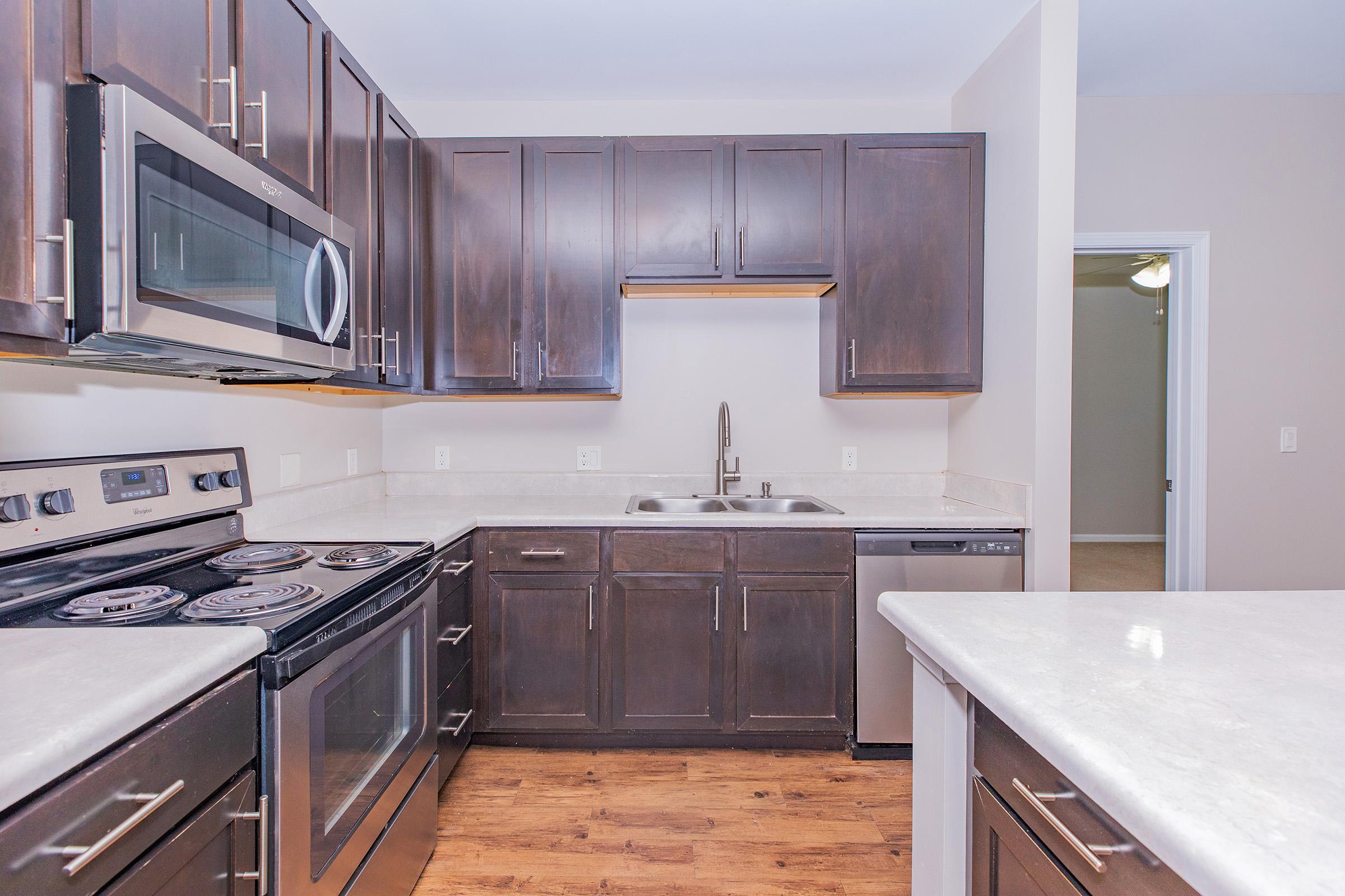 Modern kitchen featuring dark wood cabinets, stainless steel appliances, and a compact design. The space includes a microwave, oven, and dishwasher, with a double sink under a window. The countertops are light-colored, providing contrast to the cabinetry, and the flooring is a warm wood tone.