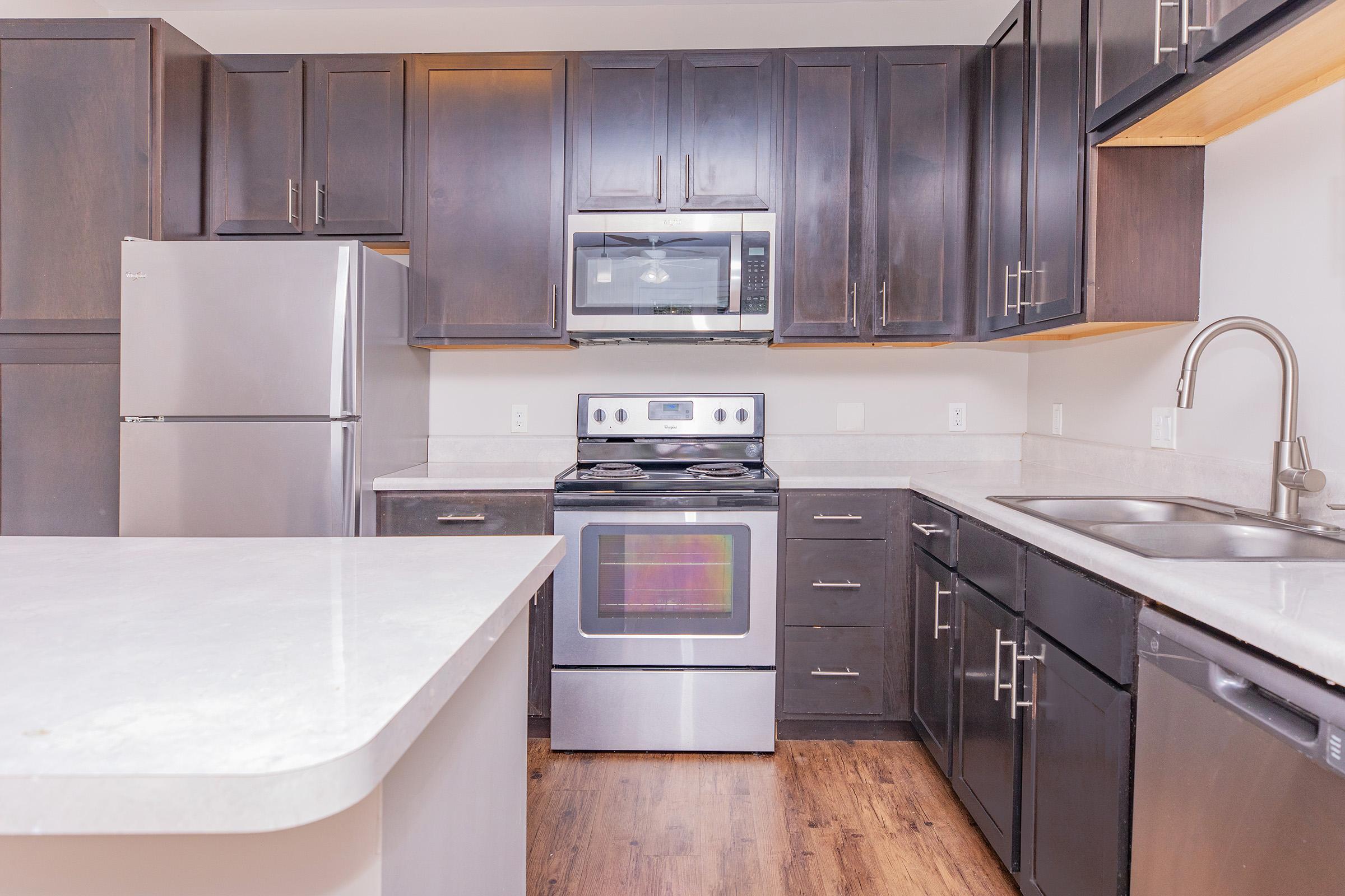 Modern kitchen featuring dark wooden cabinets, stainless steel appliances including a refrigerator, microwave, and oven. The countertops are light and smooth, with a double sink visible. The kitchen has an open layout with ample natural light, creating a spacious and inviting atmosphere.
