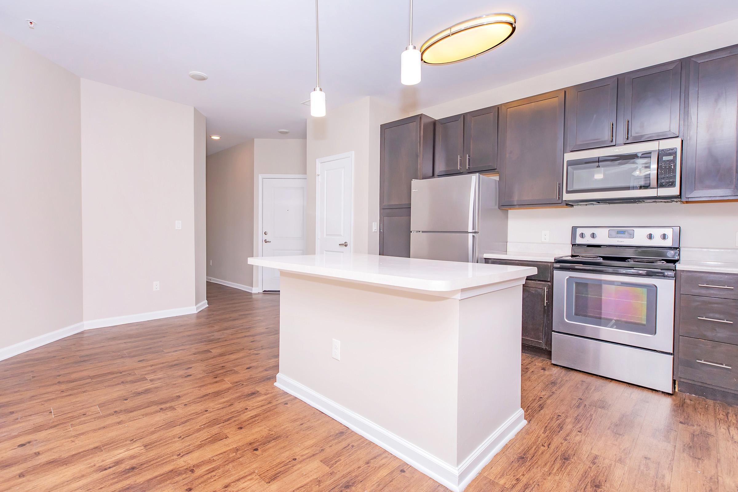 Modern kitchen featuring dark wood cabinetry, stainless steel appliances, and a center island with seating. Warm-toned hardwood flooring complements the light-colored walls. The space is well-lit with pendant lighting and has an open layout leading to a door on the left, suggesting access to another room.