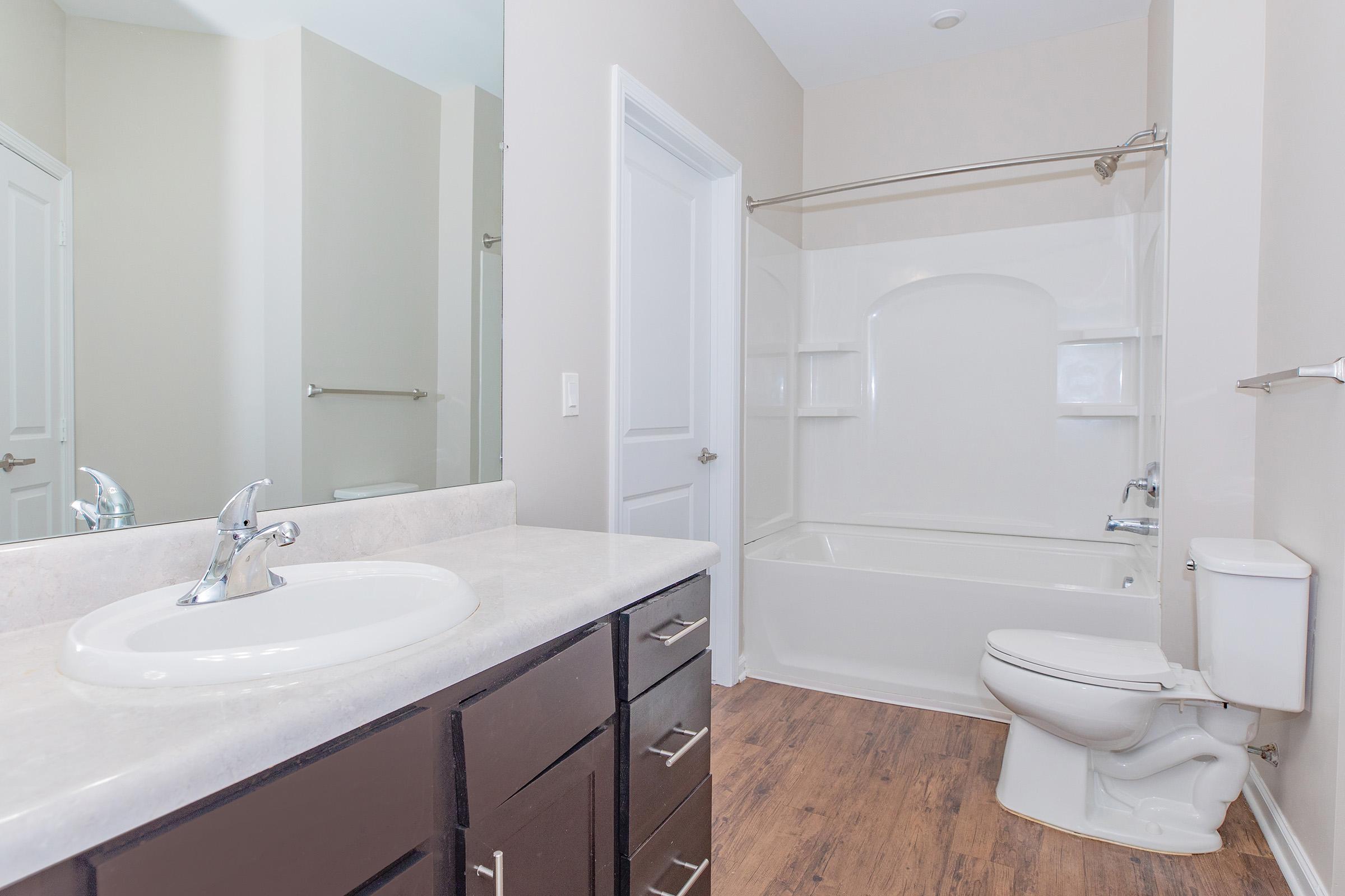 Bright and modern bathroom featuring a beige color scheme. Includes a white bathtub with a showerhead, a toilet, and a sink with a sleek faucet. The countertop is light-colored, and the cabinets are dark wood. Natural light illuminates the space, enhancing its clean and spacious feel.