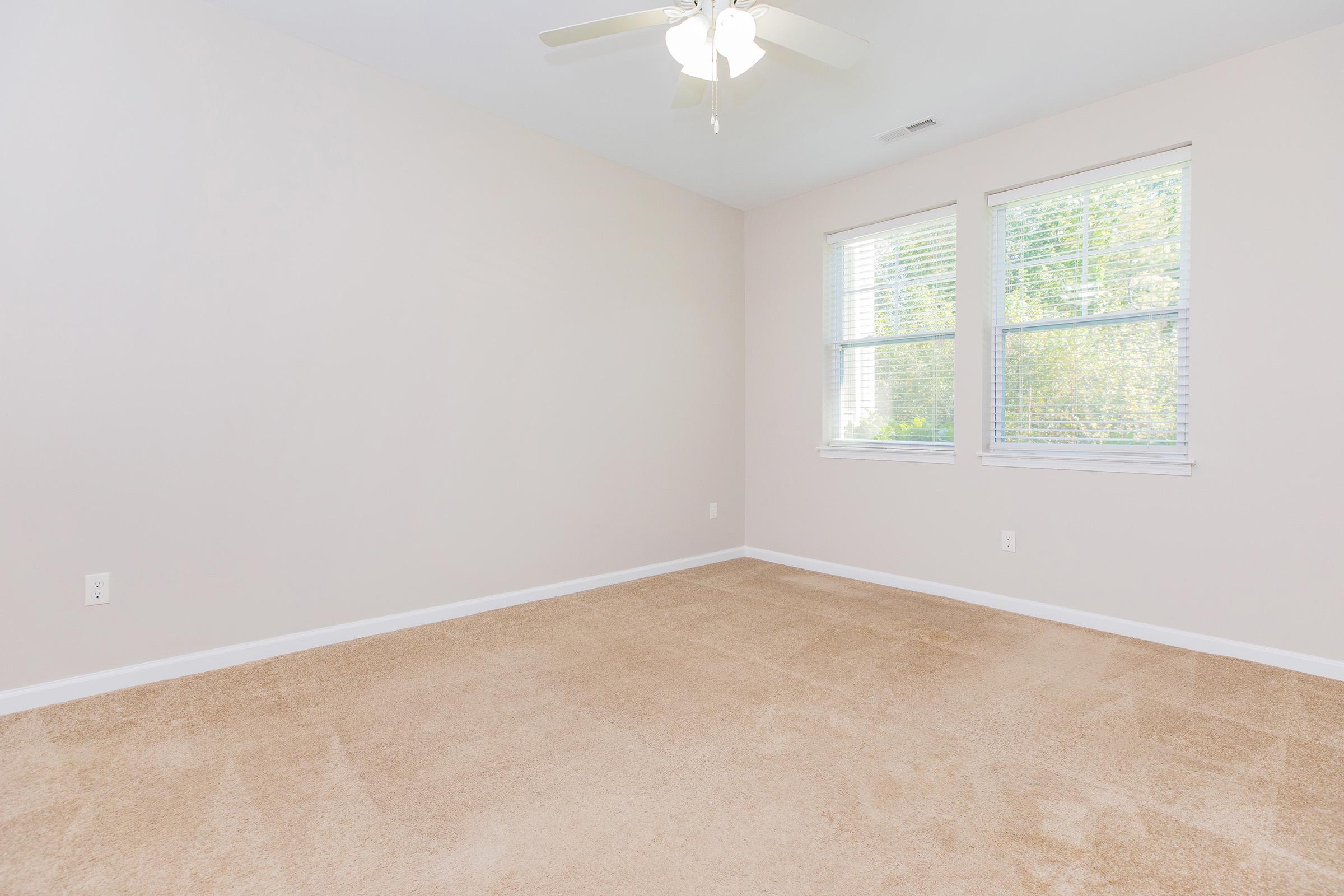 A spacious, empty room with light beige carpet and pale beige walls. Two windows with white blinds allow natural light in, and a ceiling fan is visible. The room is unfurnished, offering a clean and simple aesthetic.