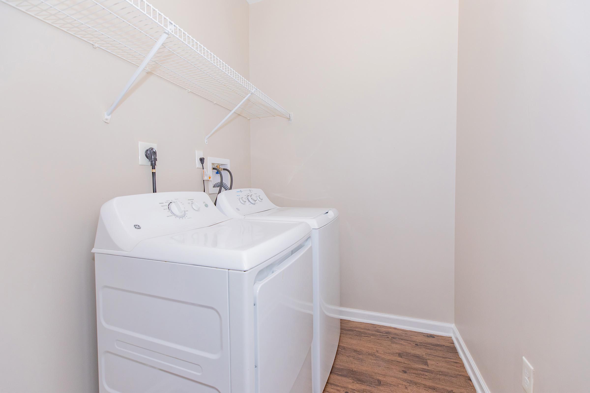 A clean laundry room featuring a white washing machine and dryer side by side against a light-colored wall. A wire shelf is mounted above the appliances, providing additional storage space. The floor is wooden, adding a touch of warmth to the space. The overall atmosphere is organized and functional.