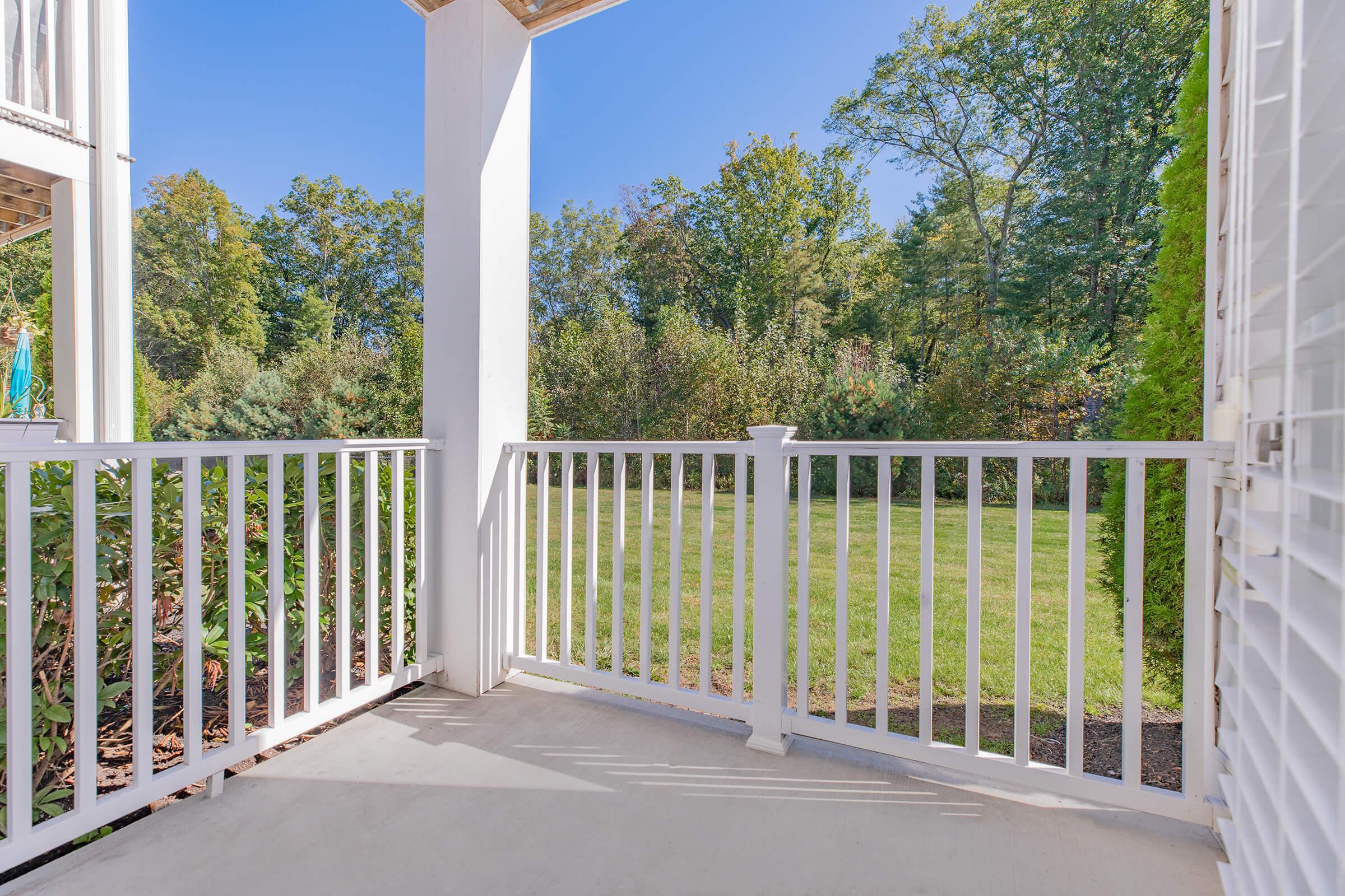 A view from a white balcony railing overlooking a green lawn and trees, with bright blue sky above. The space is well-lit, showcasing a serene outdoor environment.