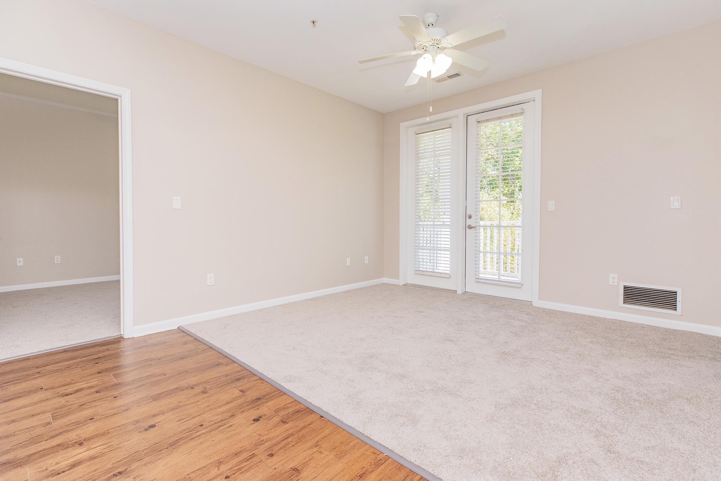 A spacious, light-filled living room with beige walls and carpet. There are two doorways leading to adjacent rooms, one of which has large windows and a sliding glass door opening to a balcony. A ceiling fan is installed in the center of the room, adding to the airy feel.