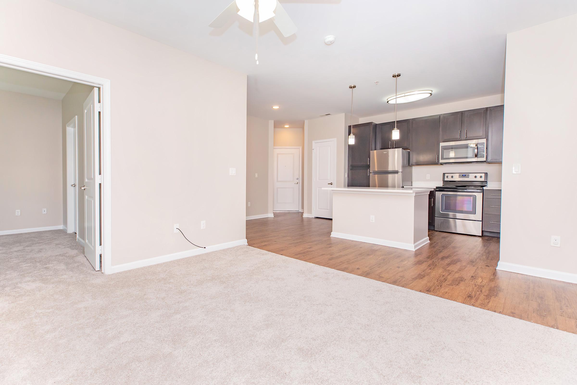A spacious, modern apartment interior featuring a living area with light-colored carpet, a kitchen with stainless steel appliances and a large island. The walls are neutral, and there are white doors leading to other rooms. Natural light brightens the space from windows.