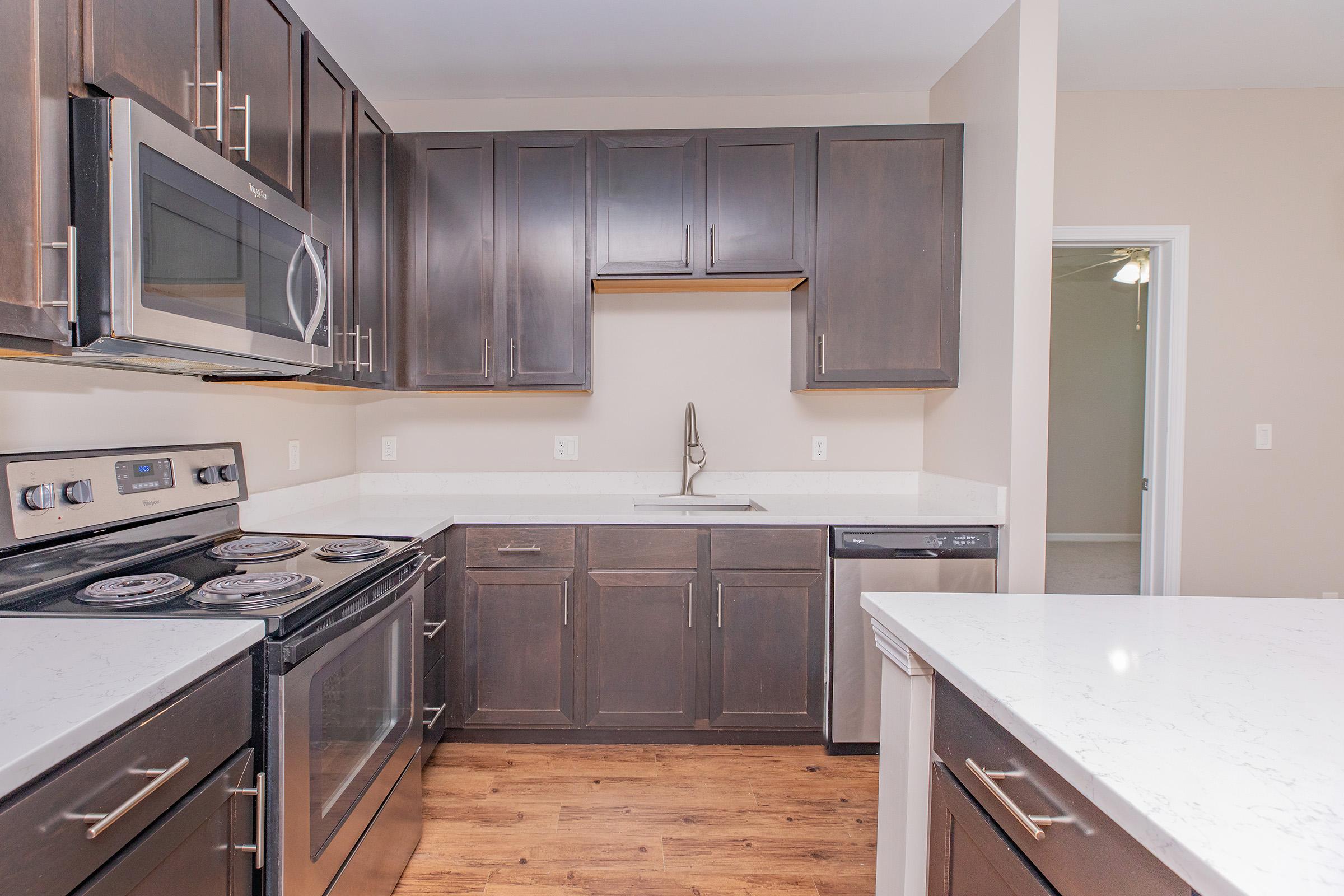 Modern kitchen featuring dark cabinetry, stainless steel appliances, including an oven, microwave, and stovetop. The countertops are white with a subtle pattern, complemented by a light-colored backsplash. A spacious layout includes a double sink and dishwasher, with hardwood flooring visible.