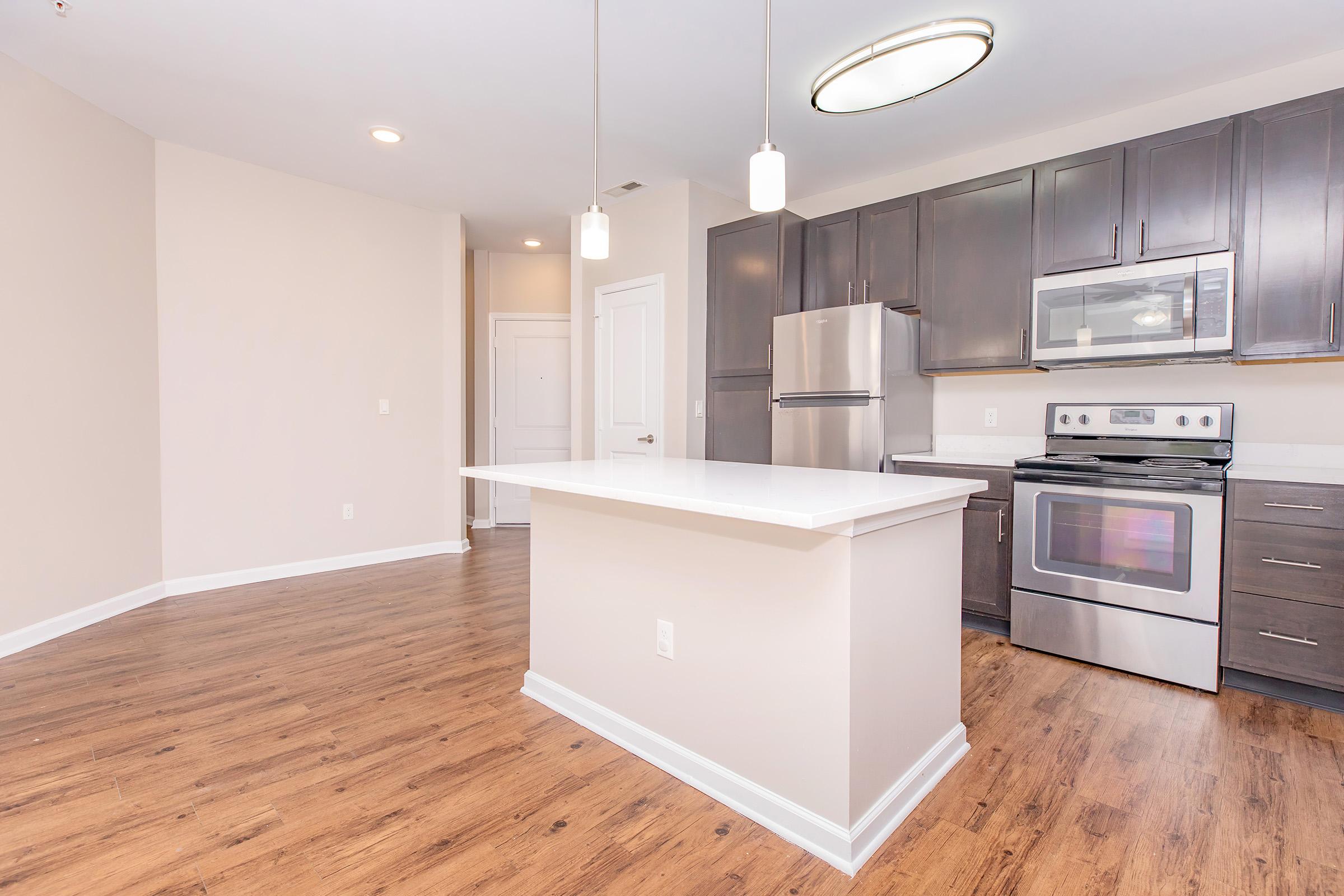 A modern kitchen featuring dark wood cabinets, stainless steel appliances, and a central island with a white countertop. The space has hardwood floors, recessed lighting, and a neutral color scheme, creating a bright and inviting atmosphere.