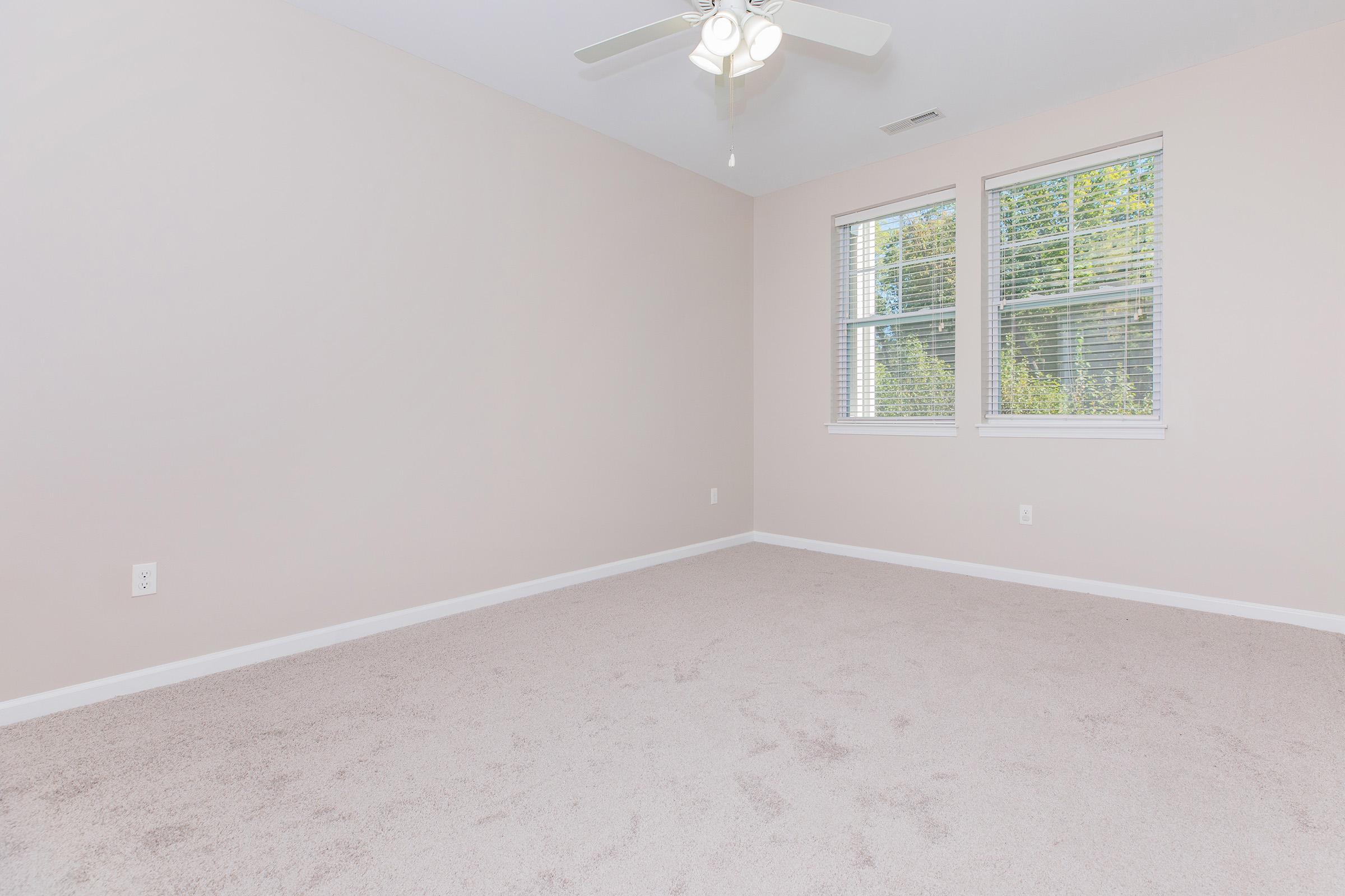 Empty room with light beige walls and carpeted flooring. A ceiling fan is mounted in the center, and there are two windows allowing natural light to enter. The space is uncluttered, creating a bright and airy atmosphere, ideal for various uses like a bedroom or office.