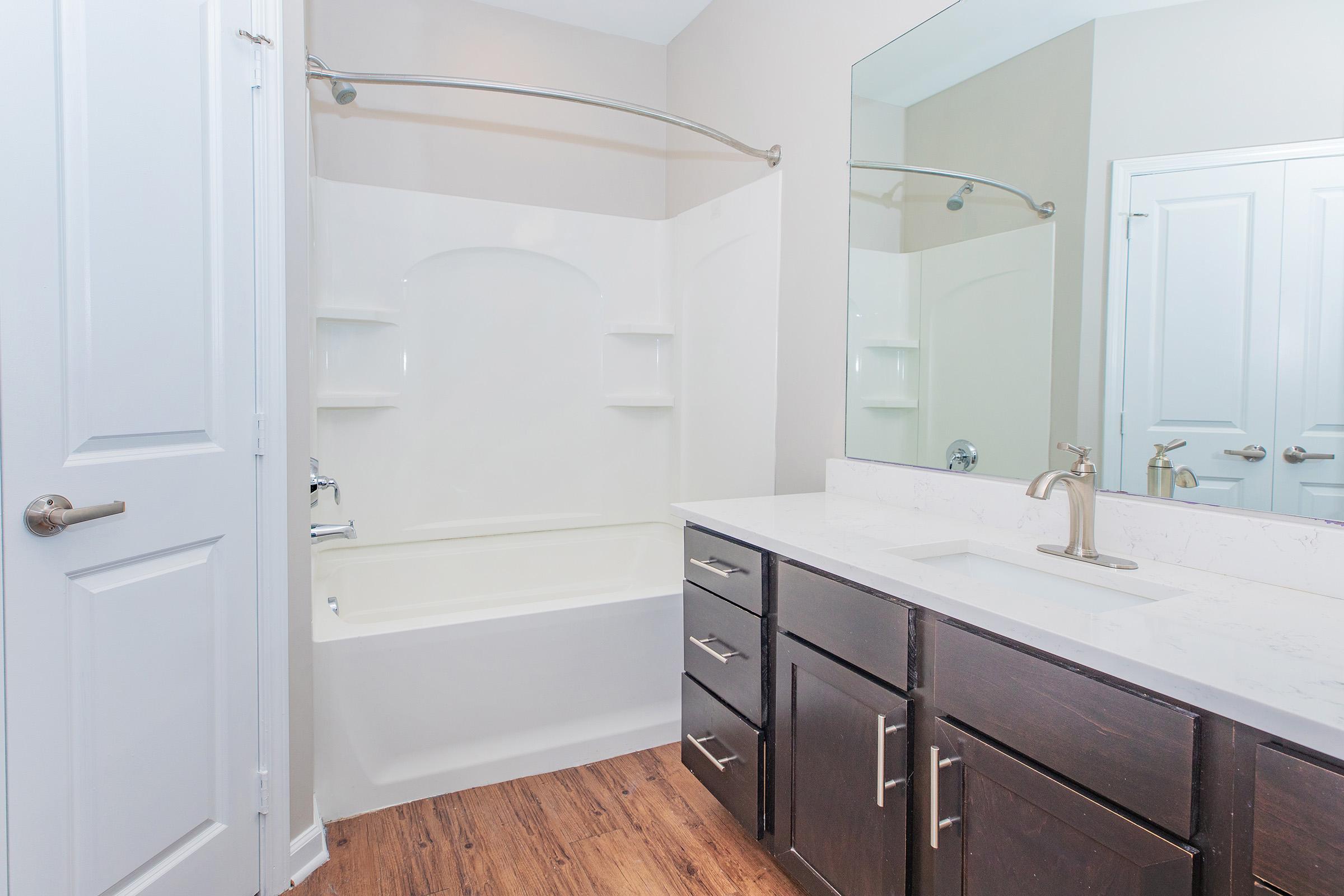 A modern bathroom featuring a white bathtub with a shower curtain, two built-in shelves, a light-colored wall, and a large mirror above a stylish dark wood vanity with a sink. The floor is covered with wood-like laminate, and there is a door leading to another room.