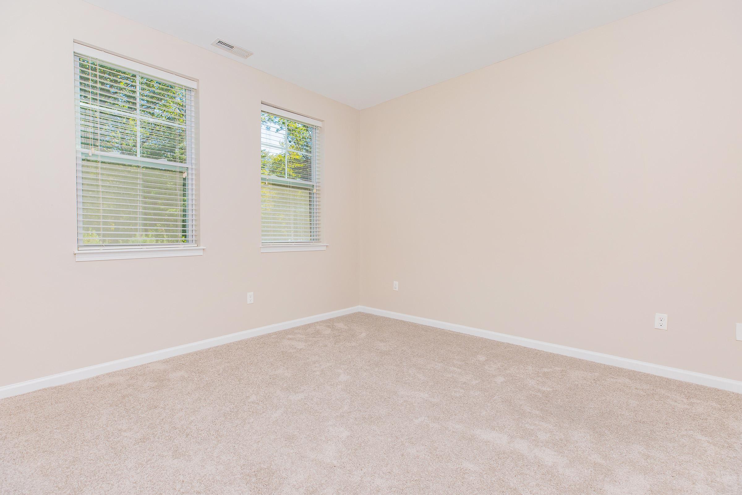 Empty room with beige walls and plush, light-colored carpet. Two windows with white blinds allow natural light to enter, illuminating the space. The room has a simple design, with no furniture or decorations present, creating a calm and spacious atmosphere.