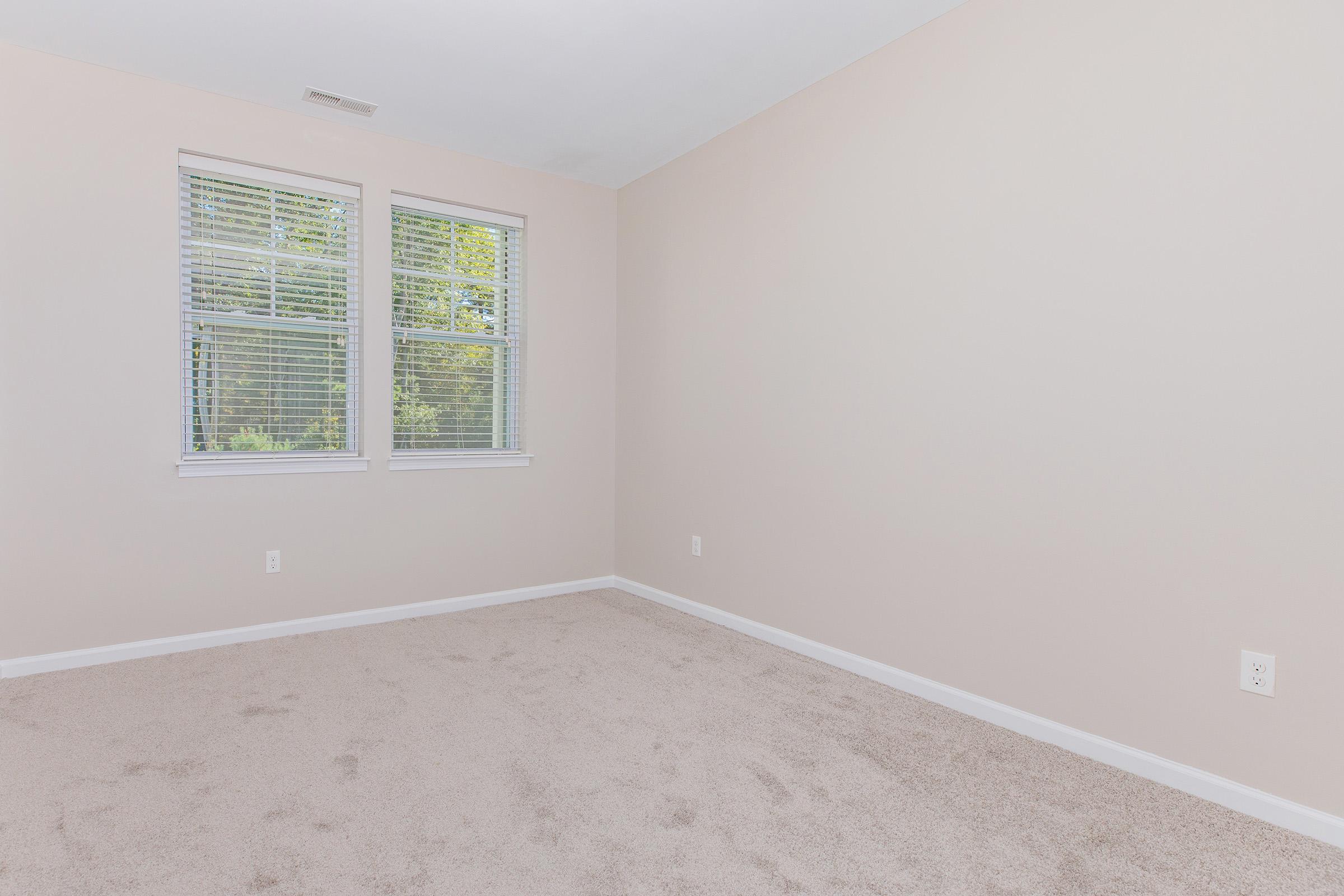 A vacant room with beige walls, plush beige carpet, and two white-framed windows that let in natural light. The space is unfurnished and tranquil, highlighting its potential for customization.