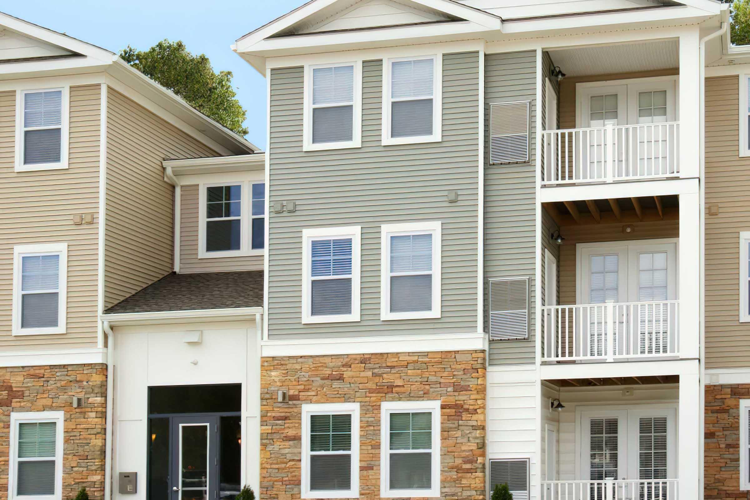 A multi-story residential building featuring a mix of light brown and green siding, stone accents, and several balconies. The entrance is framed by large windows, and the structure has a modern design with a well-maintained appearance. Trees are visible in the background.