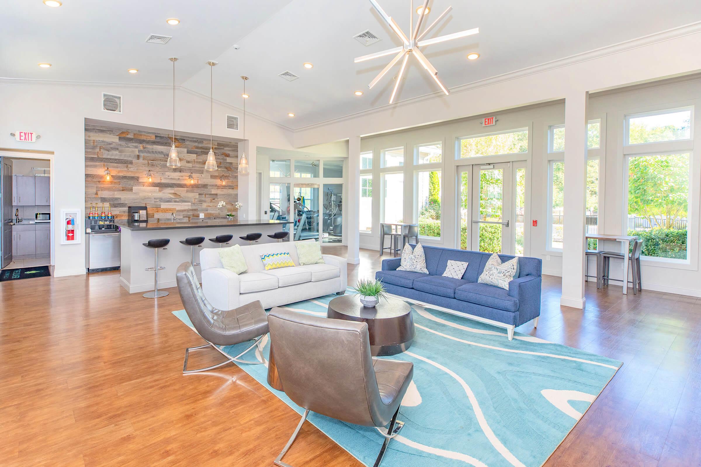 Modern living area featuring a stylish white sofa and a blue couch, surrounded by contemporary chairs. A wooden bar area with stools is in the background, and large windows allow natural light to fill the space. Decorative plants and a colorful area rug enhance the inviting atmosphere.
