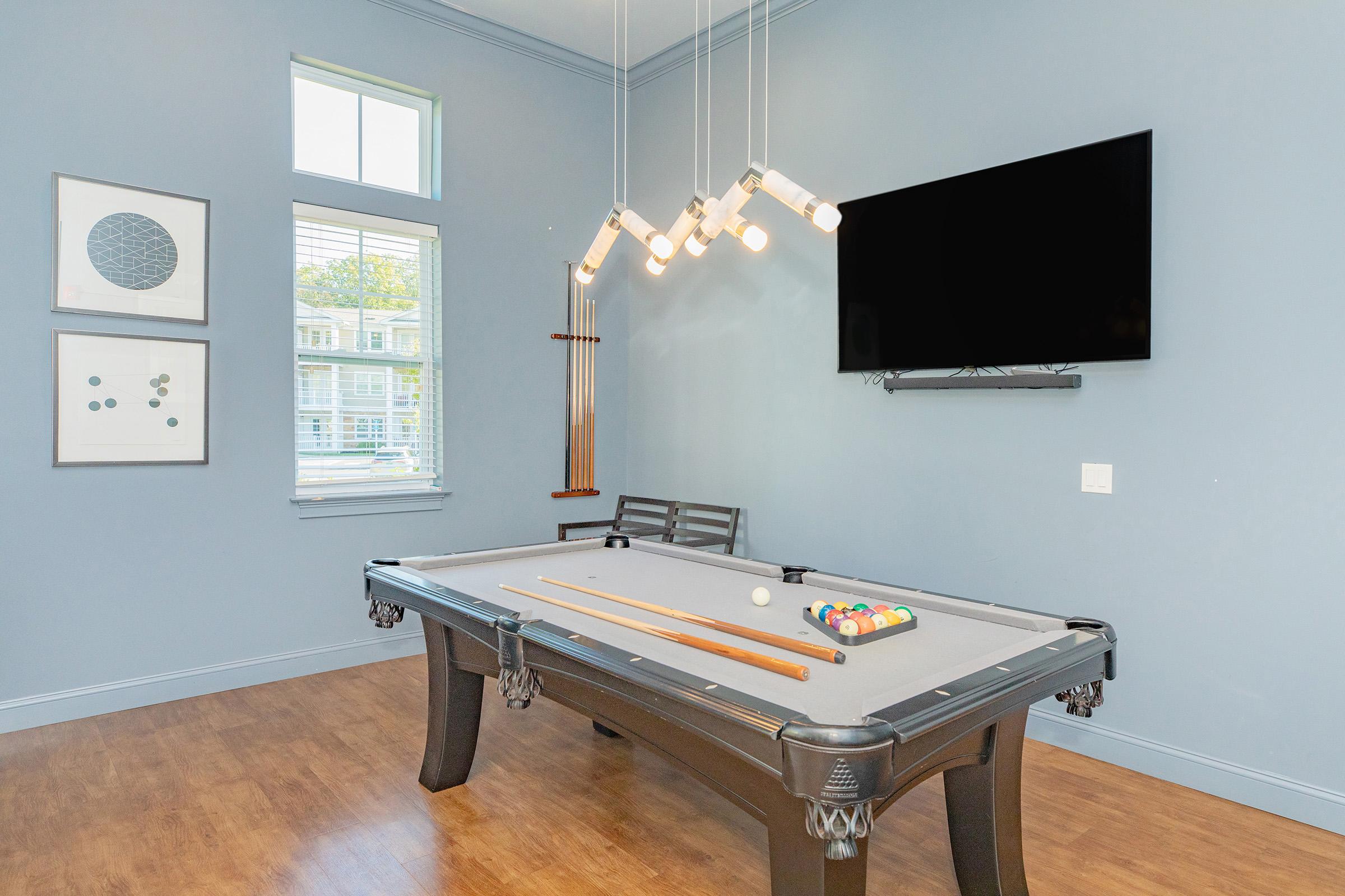A spacious game room featuring a billiards table with a full set of balls, complemented by a stylish light fixture above. The walls are painted a soft blue, and there are two framed artworks on the left. A wall-mounted television is positioned above the table, with a window providing natural light.