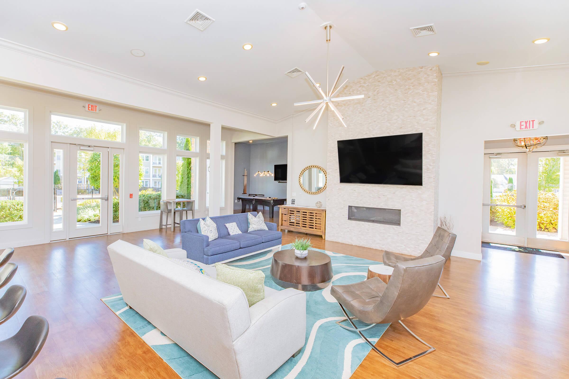 A modern living room with a light color scheme featuring a blue sofa, a white sofa, and two stylish chairs. A large flat-screen TV is mounted on the wall above a decorative fireplace. A round coffee table sits on a patterned rug, and large windows allow natural light to illuminate the space.