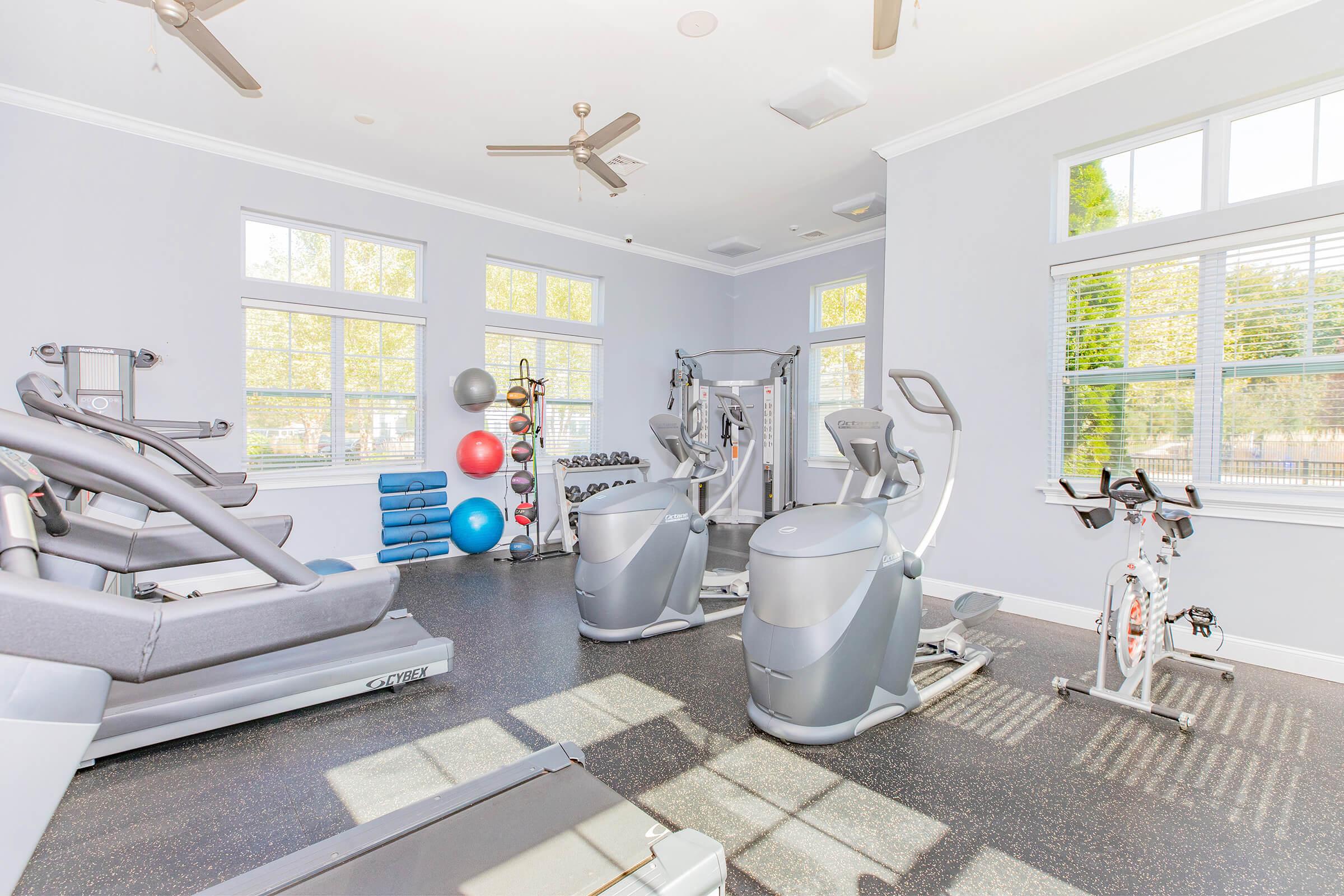 A bright and spacious gym with multiple exercise machines, including treadmills and stationary bikes. The room features large windows allowing natural light, and is equipped with colorful exercise balls and stacked mats. The flooring is dark, providing contrast to the light-colored walls.