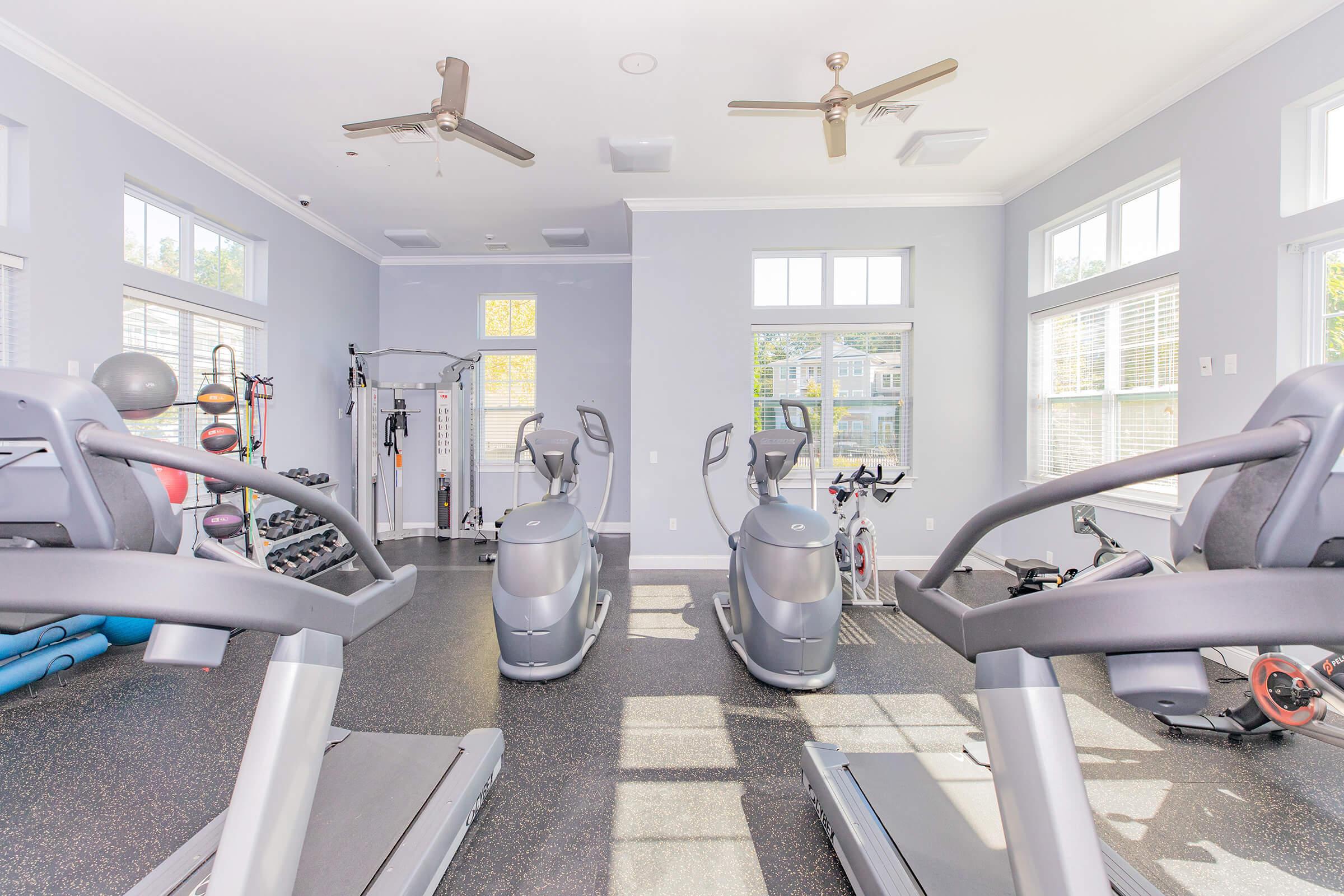 A bright and spacious gym area featuring multiple exercise machines, including treadmills and stationary bikes. The room has large windows allowing natural light, with fitness equipment like weights and a cable machine visible in the background. The flooring is dark, creating a modern and clean atmosphere.