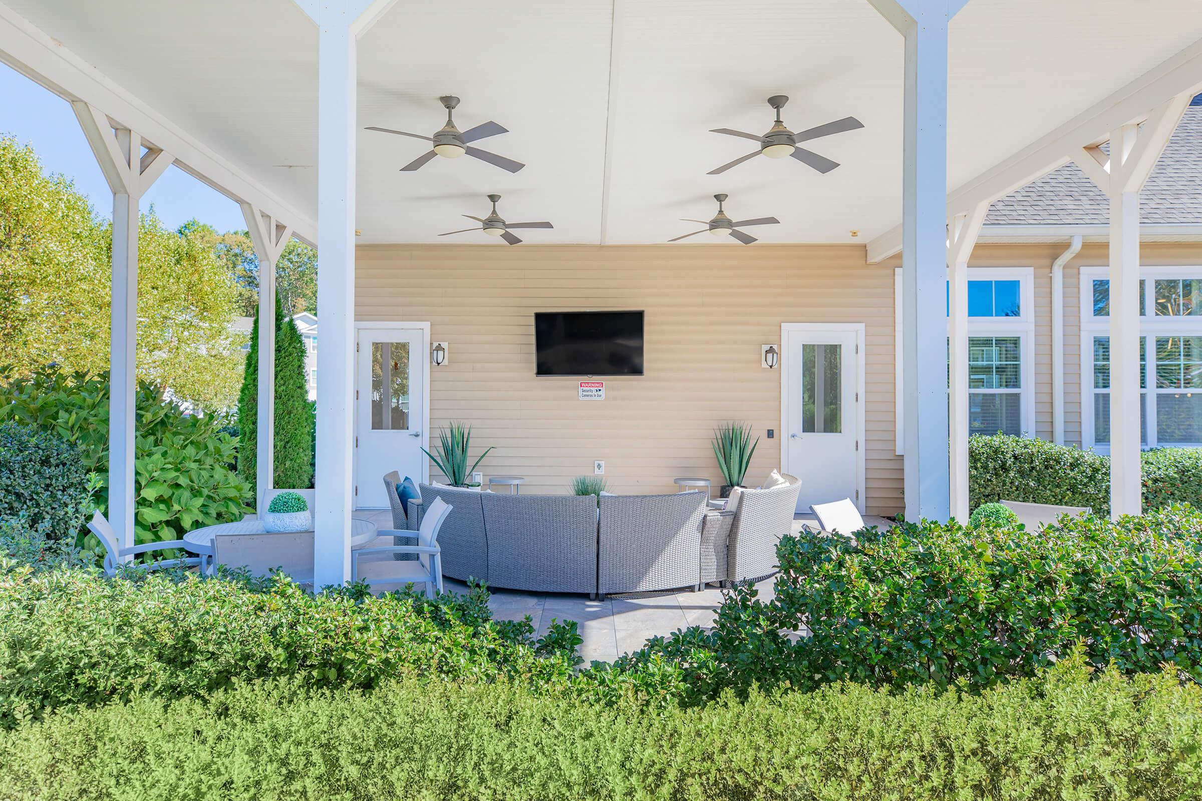 A cozy outdoor seating area featuring a gray sofa set with cushions, surrounded by greenery. Two ceiling fans are visible above, and there is a flat-screen TV mounted on the wall opposite the seating. The space is well-lit and inviting, under a covered patio structure.
