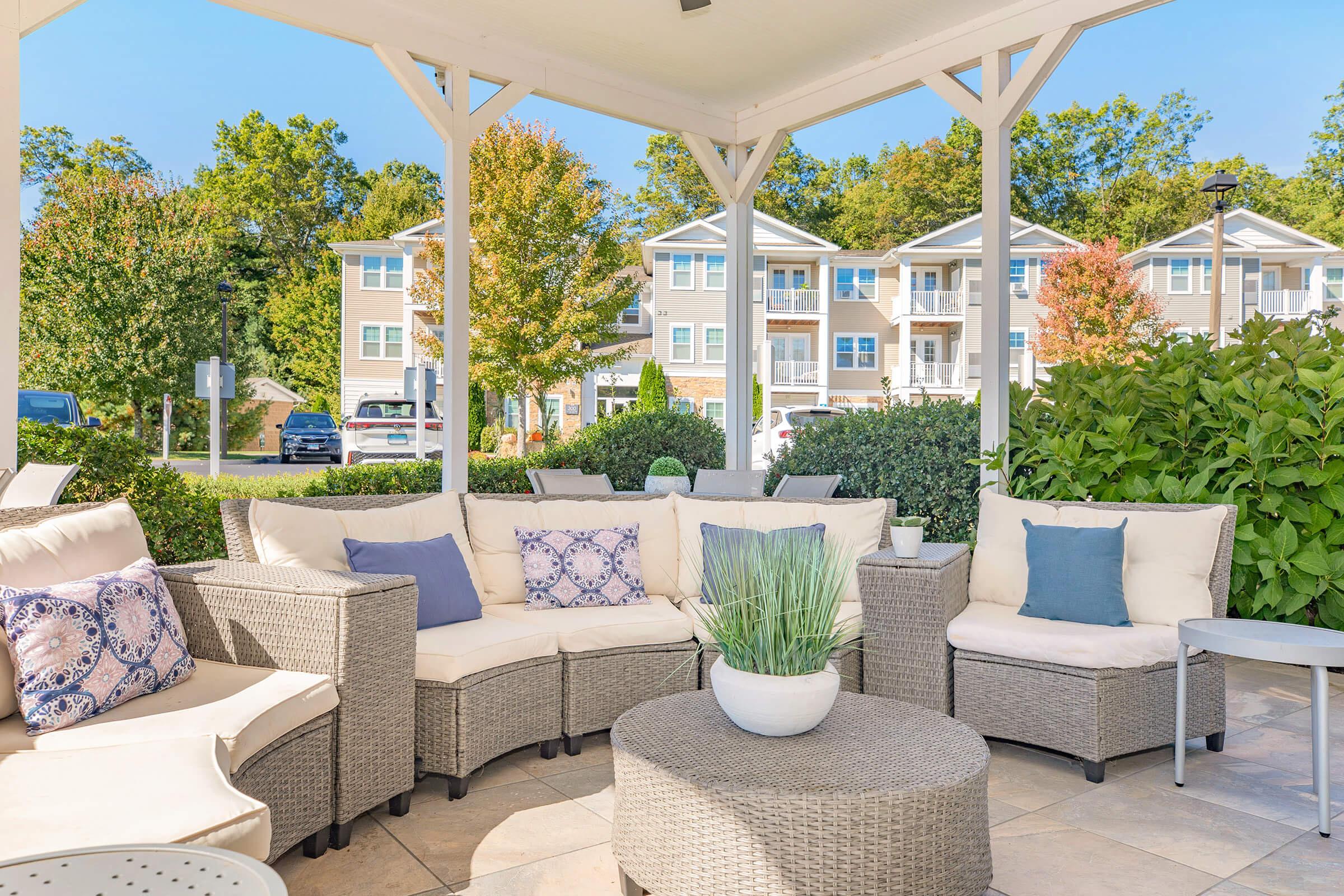 Cozy outdoor seating area with light-colored wicker couches decorated with blue and patterned cushions, arranged around a round coffee table with a potted plant. Lush green foliage surrounds the area, and modern apartment buildings are visible in the background under clear skies.