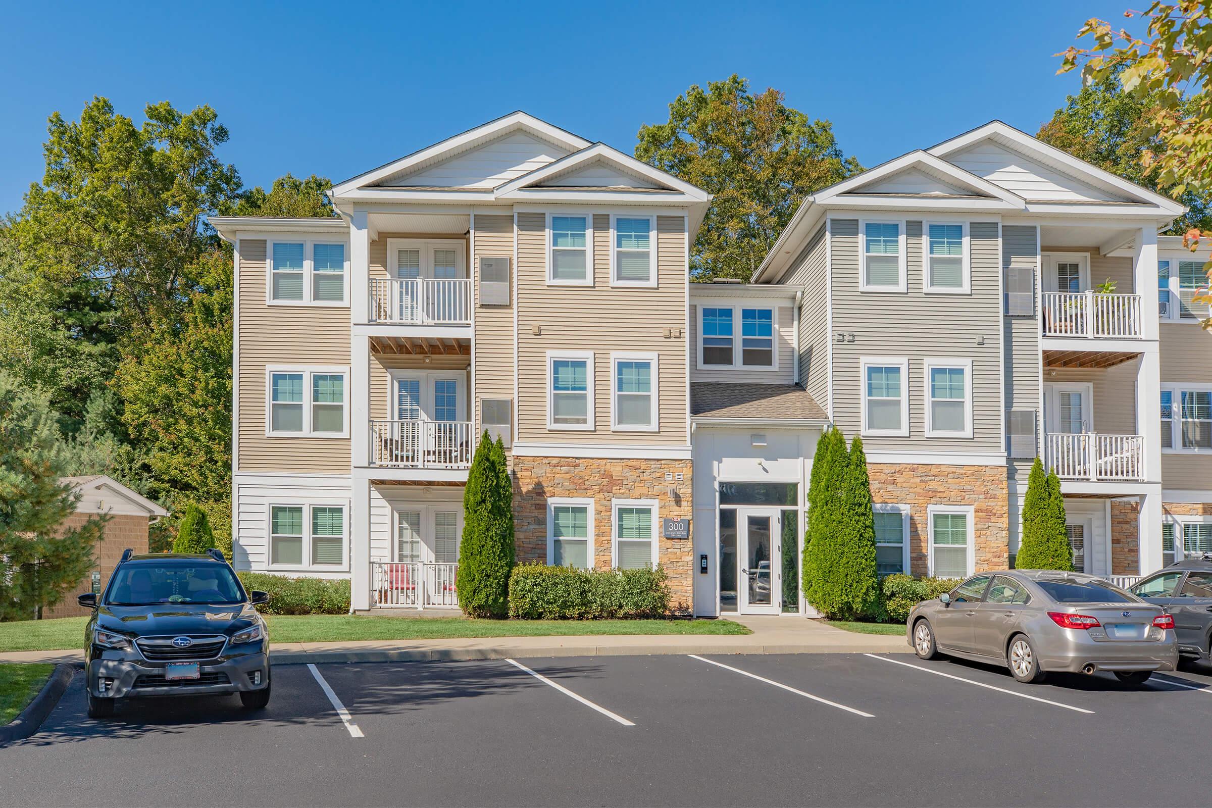 A modern apartment building with beige and gray siding, featuring balconies on the upper floors. The front includes landscaped areas with shrubs and two parked cars in the foreground. Trees are visible in the background, indicating a residential neighborhood.