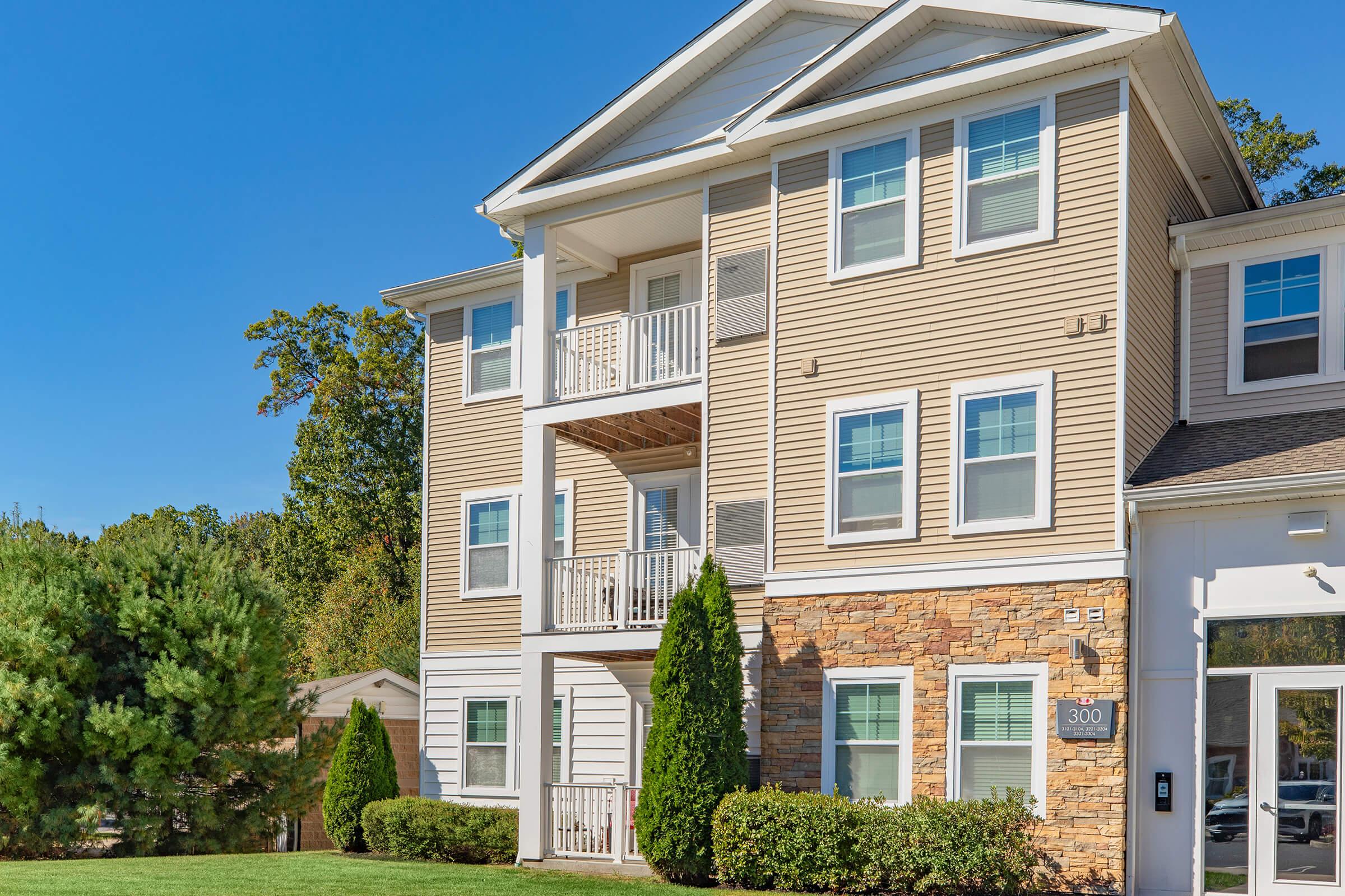 A three-story apartment building with a mix of beige siding and stone exterior. Features include several large windows, balconies on the second and third floors, and neatly maintained landscaping with shrubs and small trees. The entrance is at the ground level, marked by the number "300."