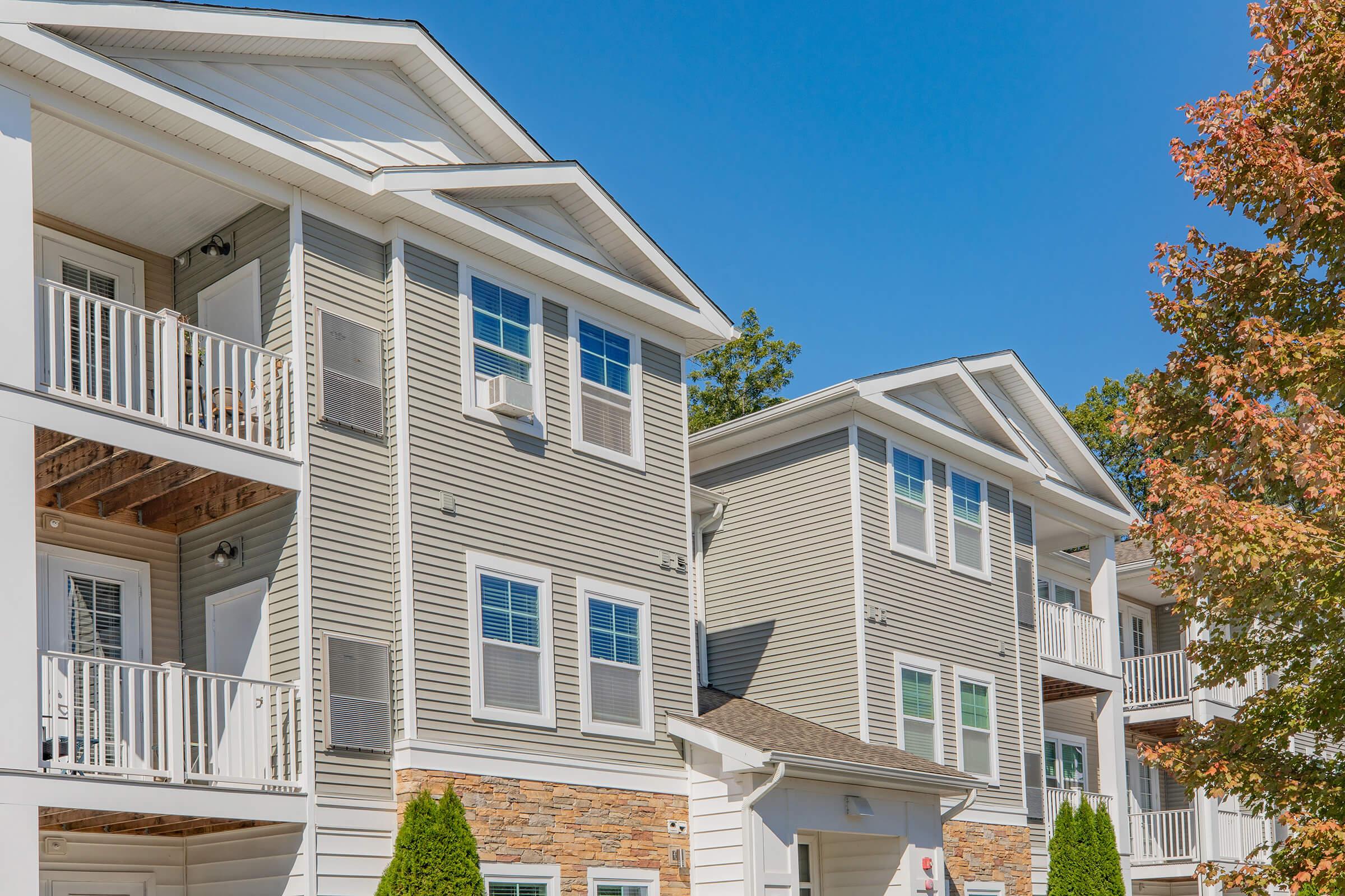 A view of modern multi-story apartment buildings with balconies, featuring a mix of beige siding and stone accents. The sky is clear and blue, with foliage from nearby trees.