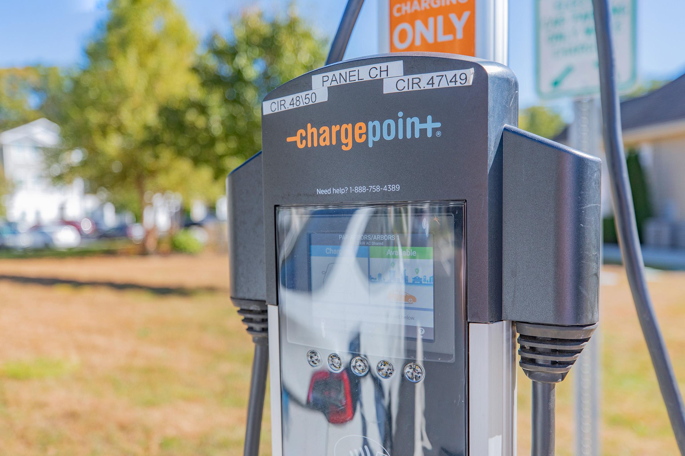 Close-up of a ChargePoint electric vehicle charging station, featuring a digital display panel labeled "PANEL CH" and nearby signage indicating "CHARGING ONLY." The background shows a grassy area with trees and buildings, emphasizing the charging station's location.