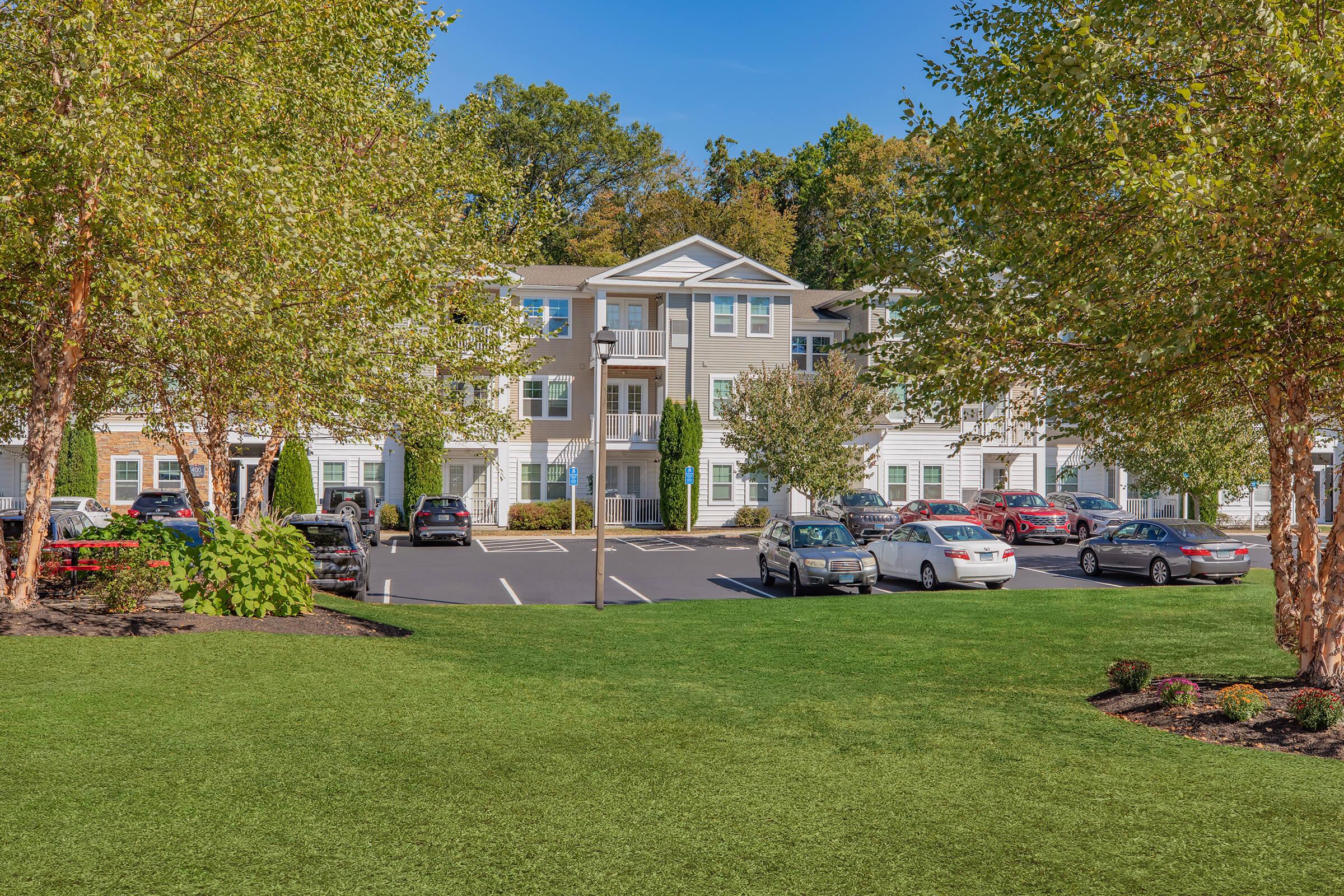 A well-maintained residential building surrounded by landscaped greenery. The image features a parking lot with several cars and trees, creating a welcoming atmosphere. The sky is clear and blue, indicating a sunny day.
