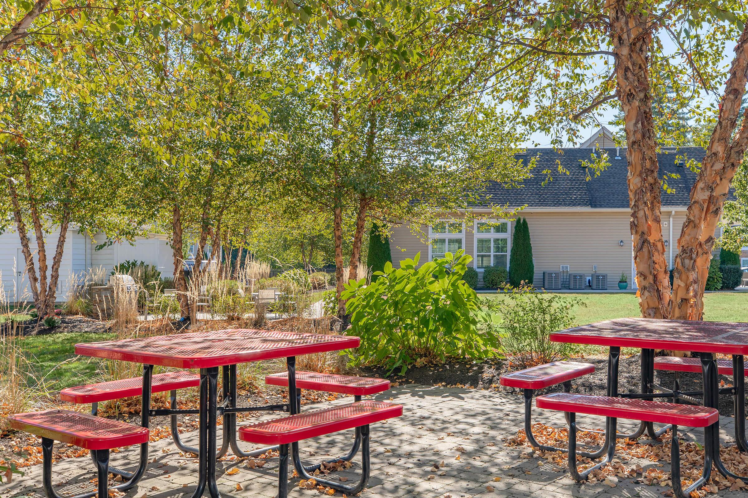 A sunny outdoor area featuring red picnic tables surrounded by lush greenery and trees. In the background, there is a light-colored building with windows and patio furniture visible. The ground is covered with leaves, indicating a seasonal change.