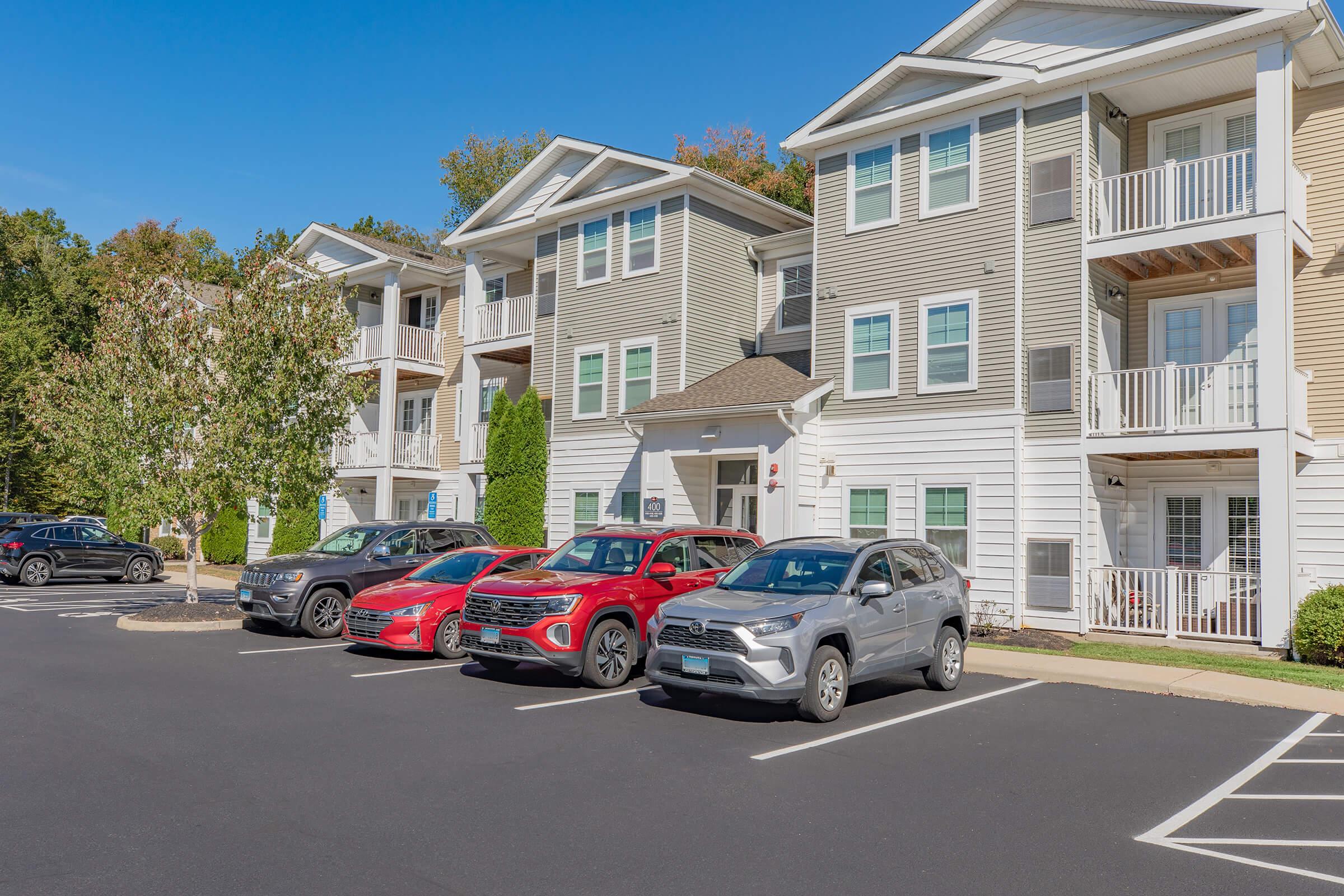 A view of a residential apartment complex featuring three buildings with balconies. There are several parked cars in the foreground, including two SUVs, a red crossover, and a silver SUV. The landscaping includes trees and shrubs, with a clear blue sky above.