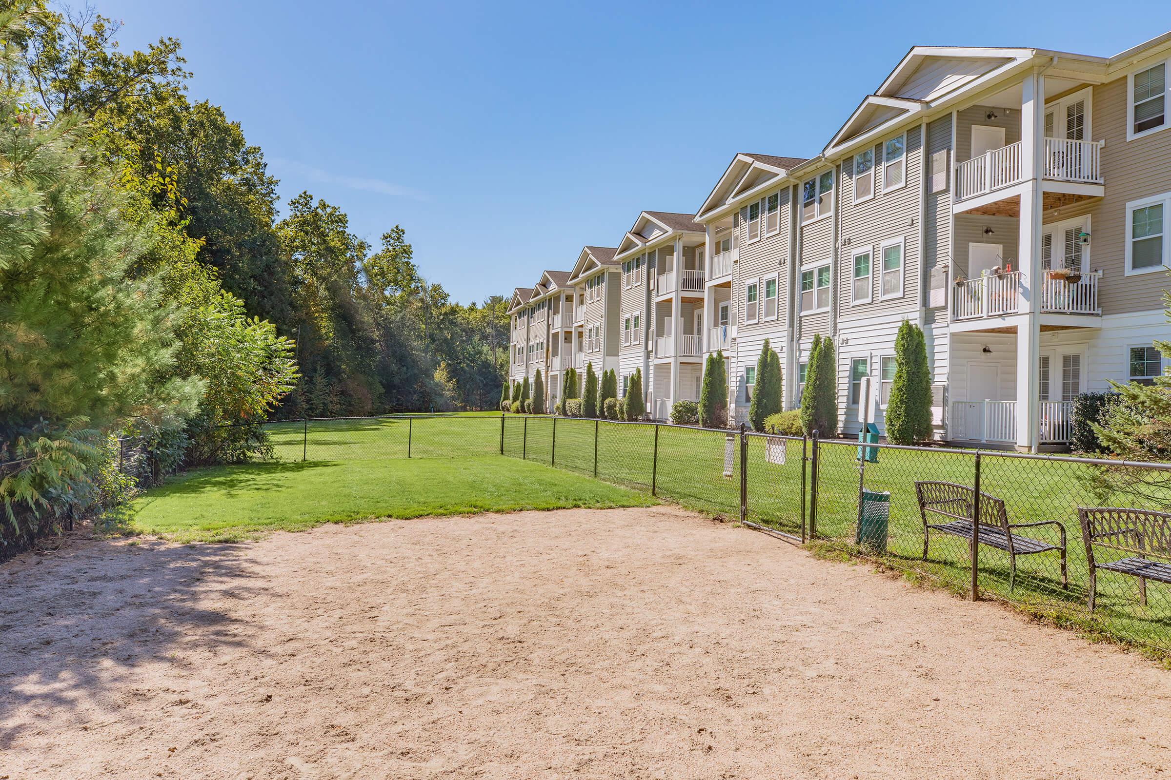 A view of a residential complex featuring three-story apartment buildings with balconies. In the foreground, there's a sandy area fenced in, possibly for a dog park, surrounded by a well-maintained lawn and trees in the background. The sky is clear and blue, creating a bright and inviting atmosphere.