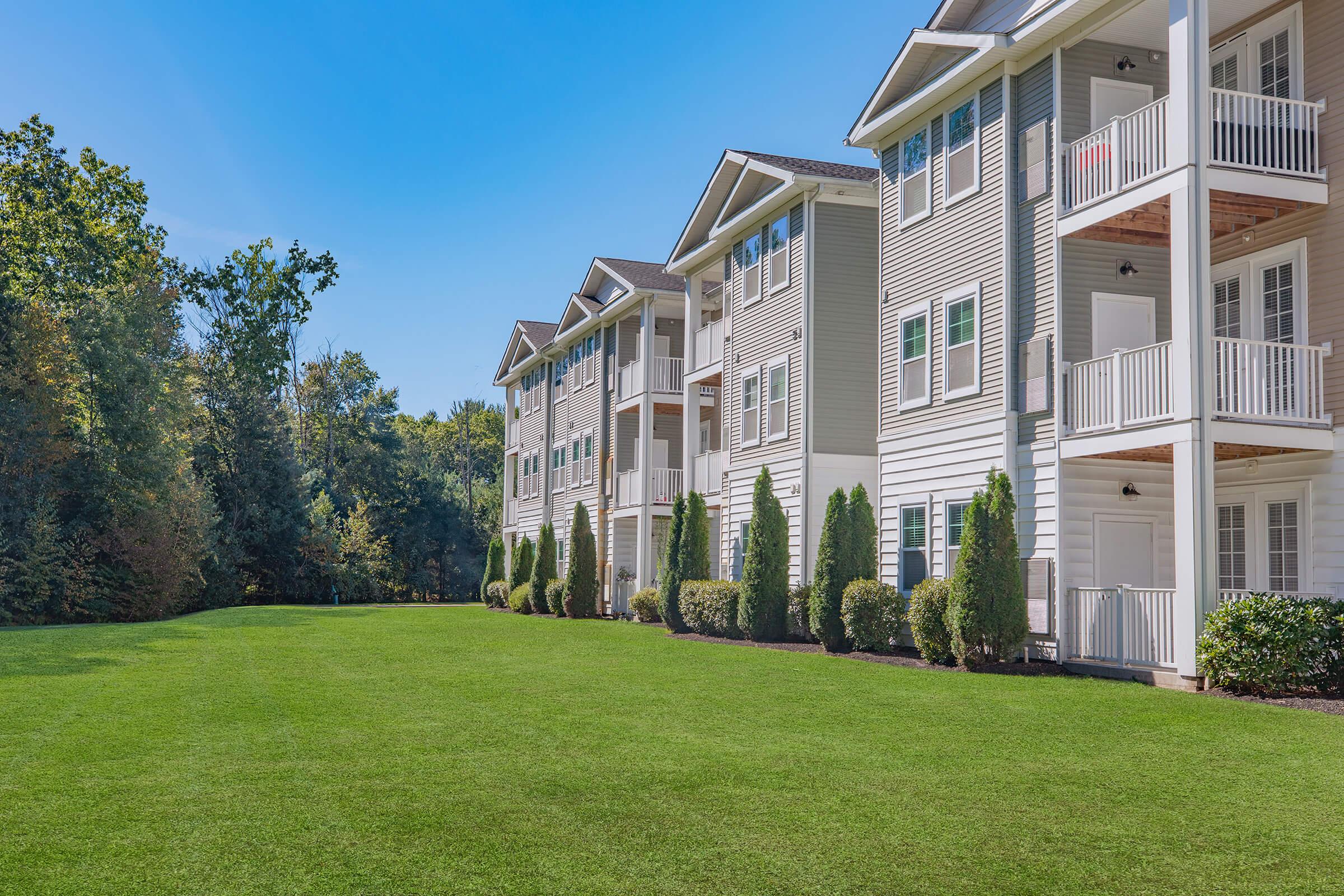 A well-maintained residential building with multiple stories, featuring balconies. The surrounding area consists of a lush green lawn and trees in the background under a clear blue sky. The scene conveys a peaceful and inviting atmosphere.