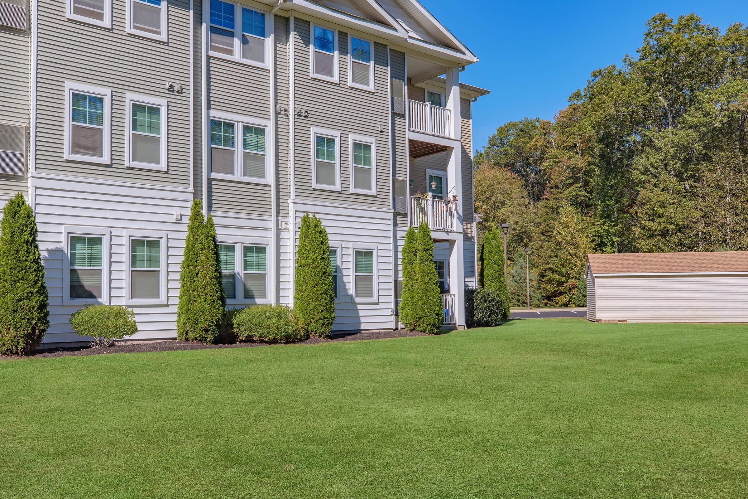 A well-maintained apartment building with a light grey exterior, featuring several windows. The grass in front is neatly trimmed and vibrant green. There are two shrubs near the building's foundation and a storage shed visible in the background among trees and a clear blue sky.