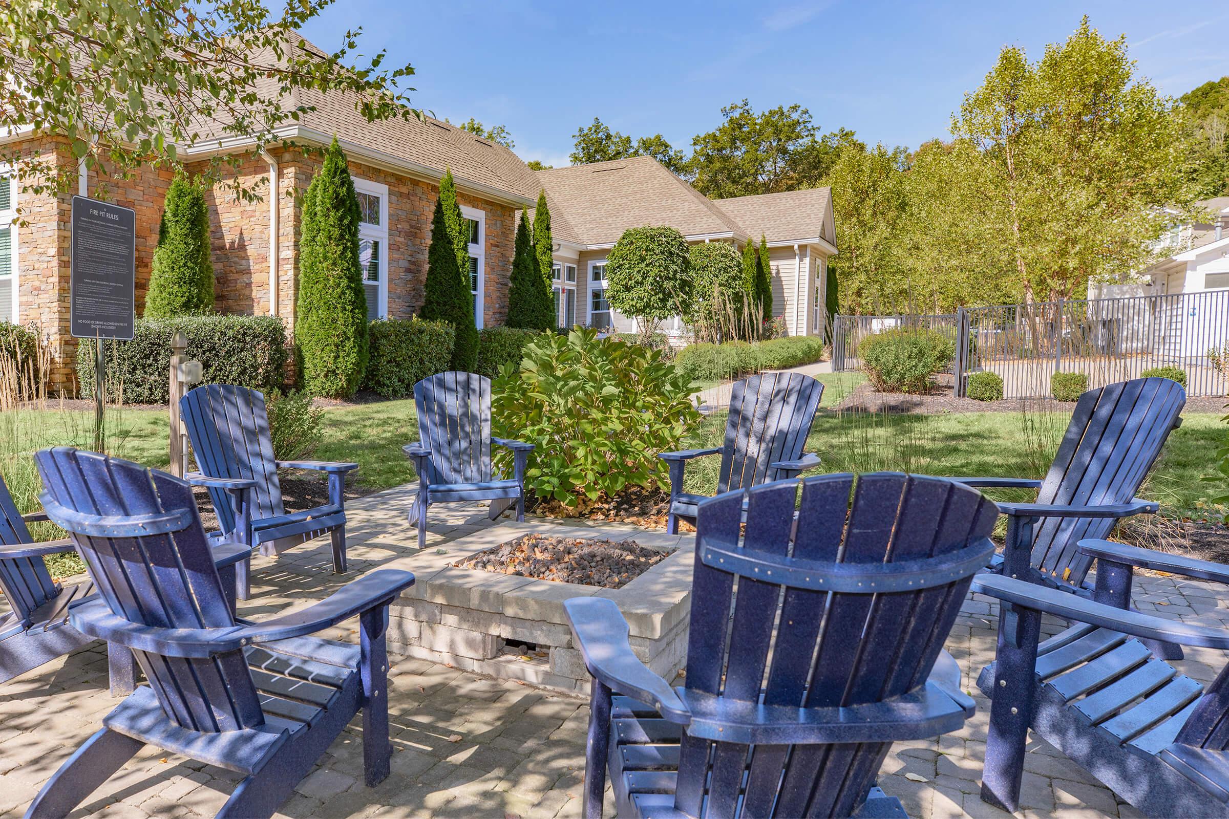 A cozy outdoor seating area featuring several blue Adirondack chairs arranged around a stone fire pit. Lush greenery and ornamental trees surround the space, with a building in the background and a clear blue sky above, creating a welcoming atmosphere for relaxation.