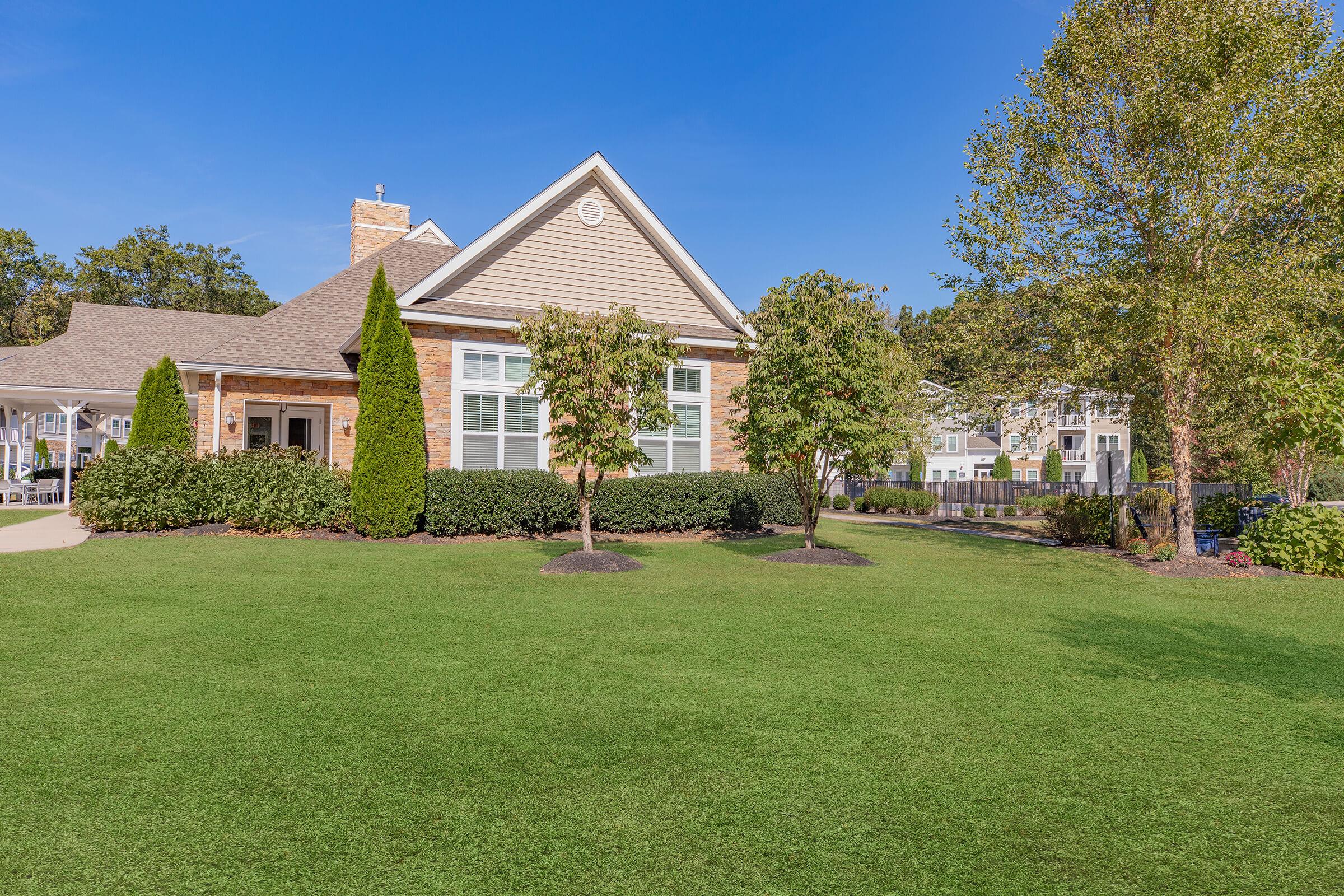 A well-maintained residential building with a landscaped front yard. The structure features a combination of brick and siding, with large windows and a gabled roof. Lush green grass surrounds the building, and there are neatly trimmed shrubs and trees, under a clear blue sky.