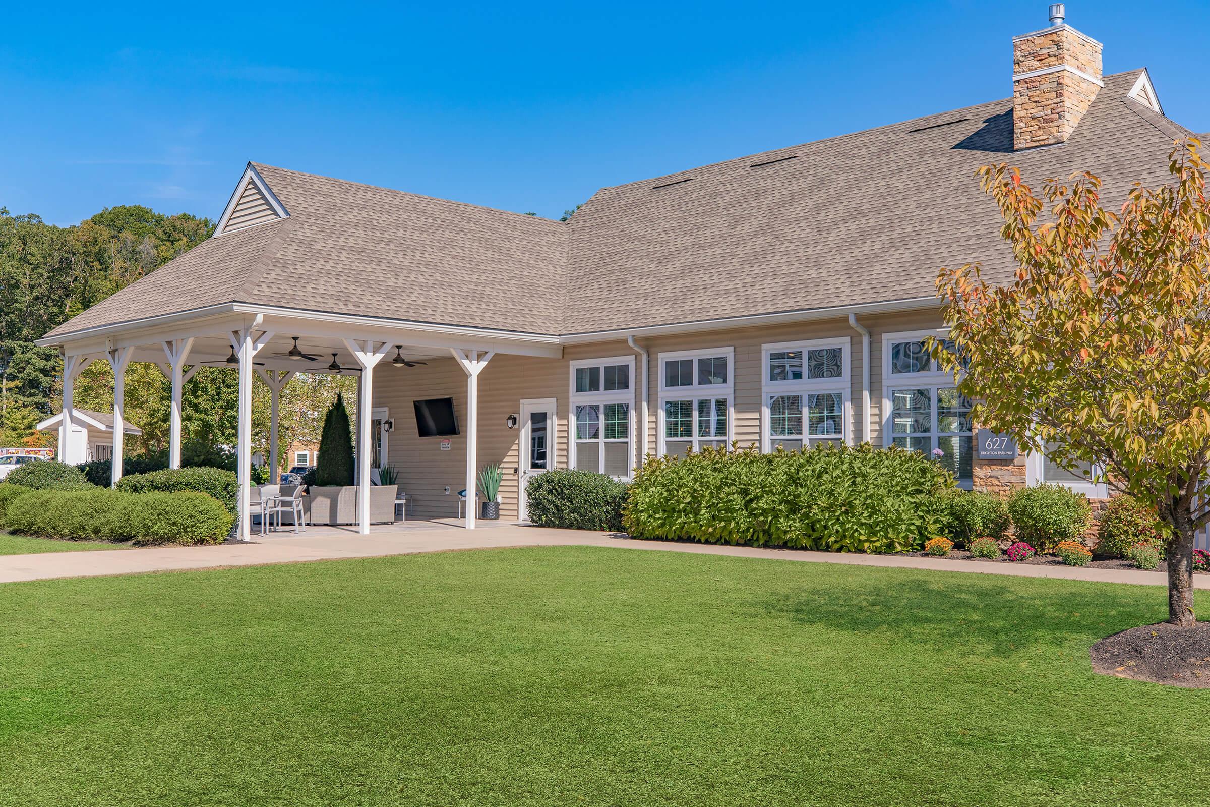 A modern, single-story building with a large front porch and multiple windows. The structure features a sloped roof, light-colored siding, and stone accents. Surrounding the building is well-maintained green grass and landscaping, including shrubs and a small tree. Clear blue sky above.