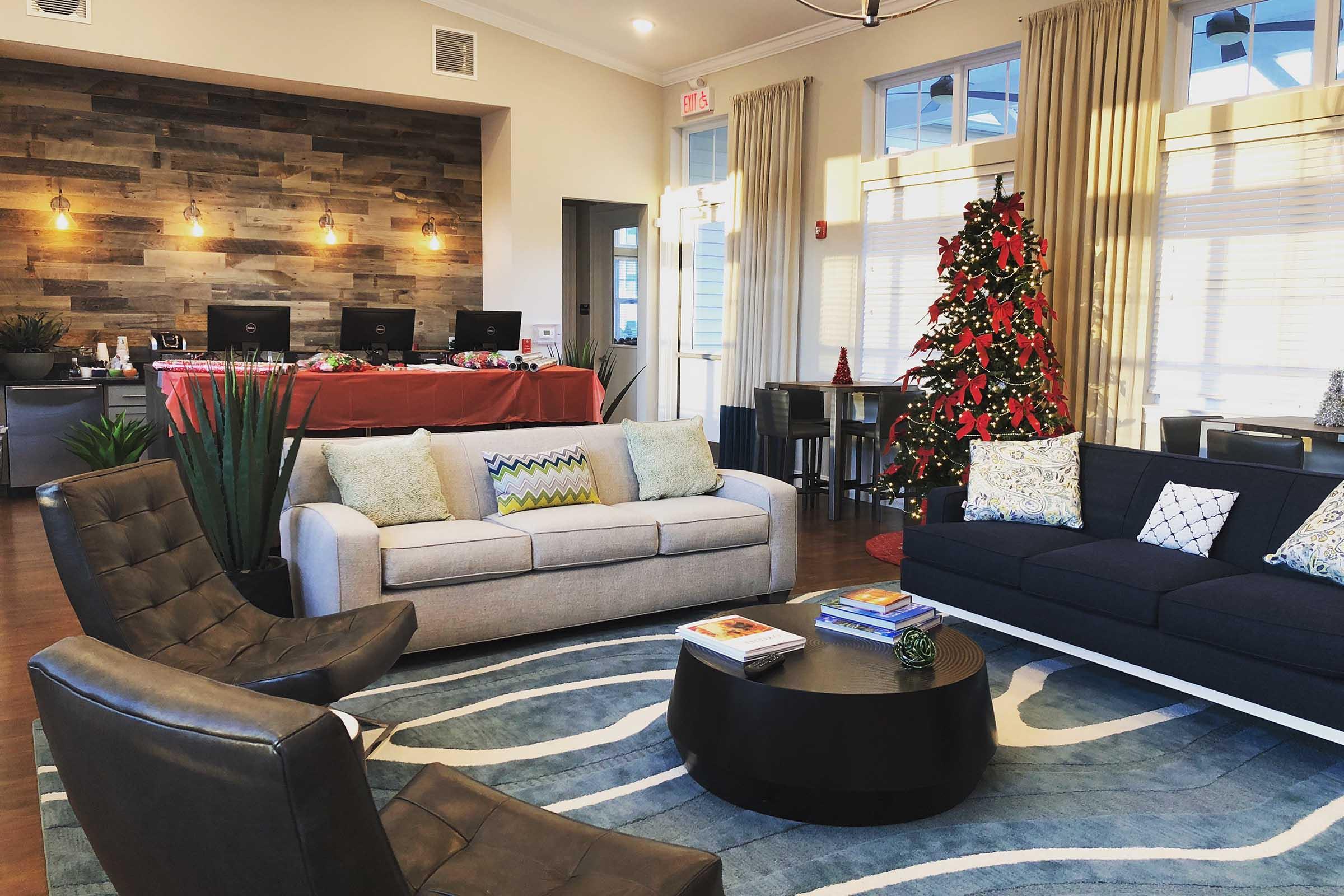 A cozy lounge area featuring a modern sofa and two stylish armchairs. A circular coffee table sits on a patterned rug, surrounded by decorative pillows. In the background, there’s a reception desk with festive decor, including a Christmas tree adorned with red ribbons and ornaments. Natural light filters through large windows.