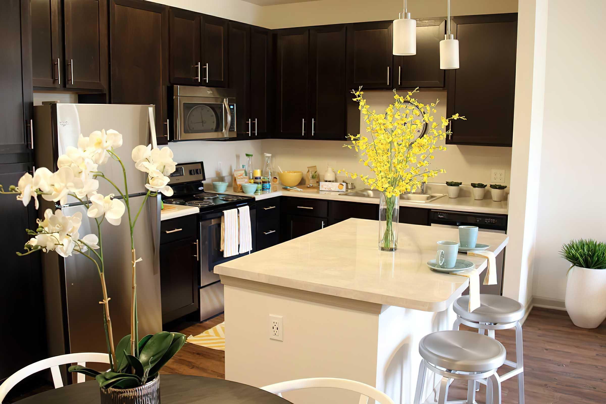 A modern kitchen featuring dark wood cabinetry and stainless steel appliances. A large island with a light countertop holds a vase of yellow flowers and two blue mugs. The dining area includes a round table with an orchid centerpiece, and there are white bar stools at the island. Bright and inviting ambiance.