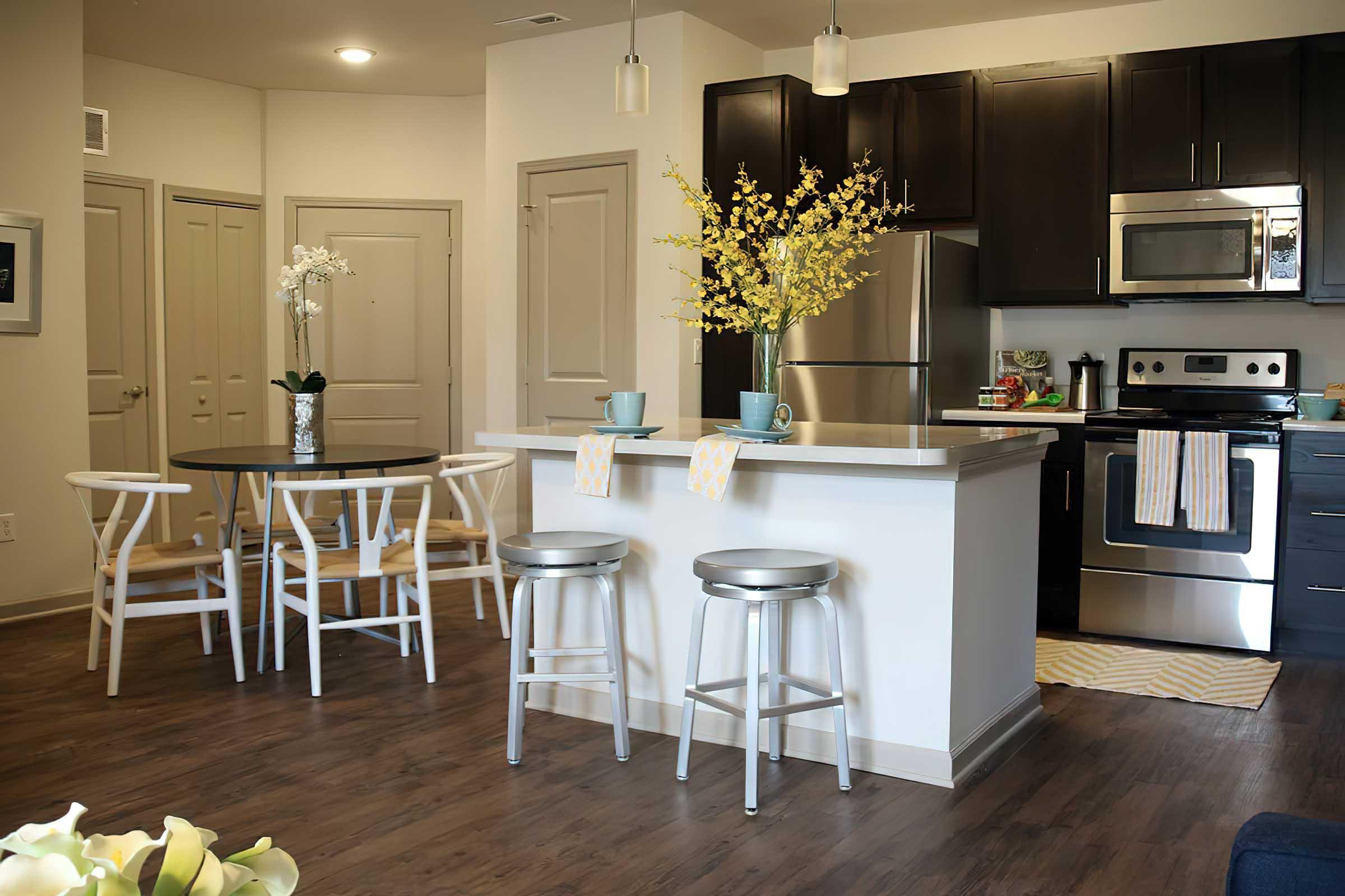 A modern kitchen and dining area featuring a large island with two bar stools, a round dining table with white chairs, stainless steel appliances, and dark cabinetry. Decorative flowers in a vase and a cozy atmosphere enhance the inviting interior design.